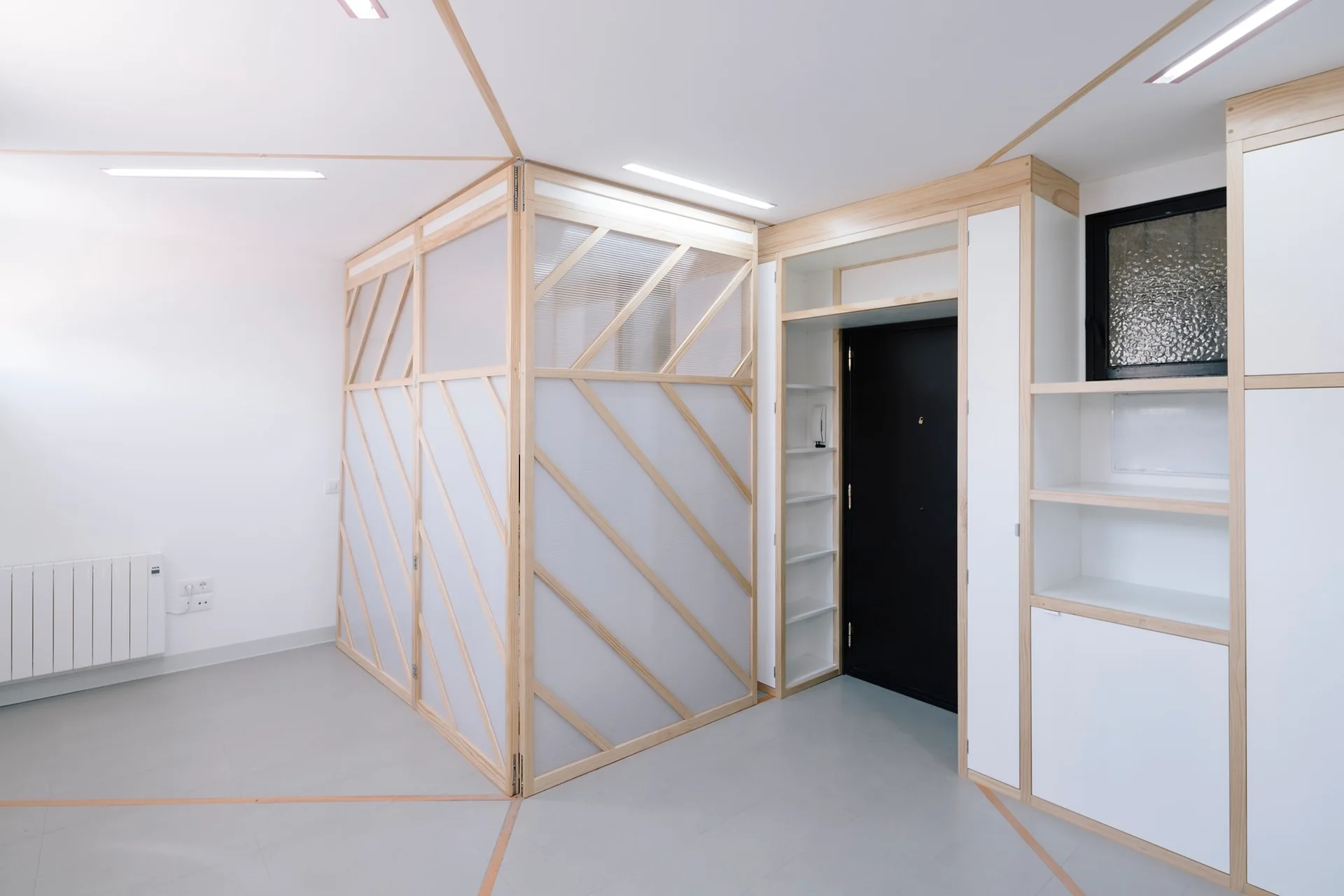 A minimalist room features a light wood and frosted glass partition with diagonal framing next to built-in white shelving and a black door.
