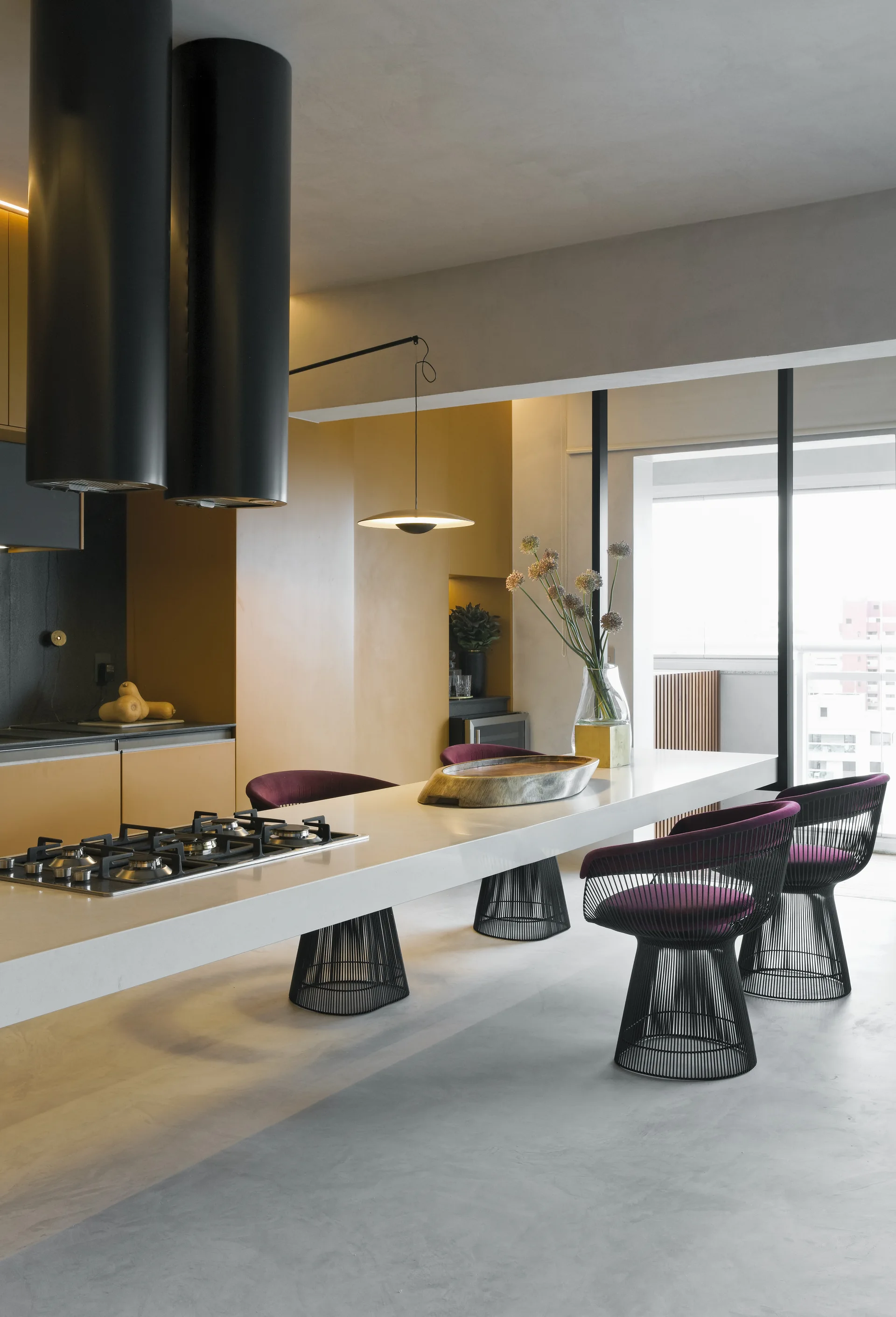 A contemporary kitchen featuring a long white island with a cooktop, black cylindrical range hoods, mustard yellow cabinets, and purple wire-frame chairs.