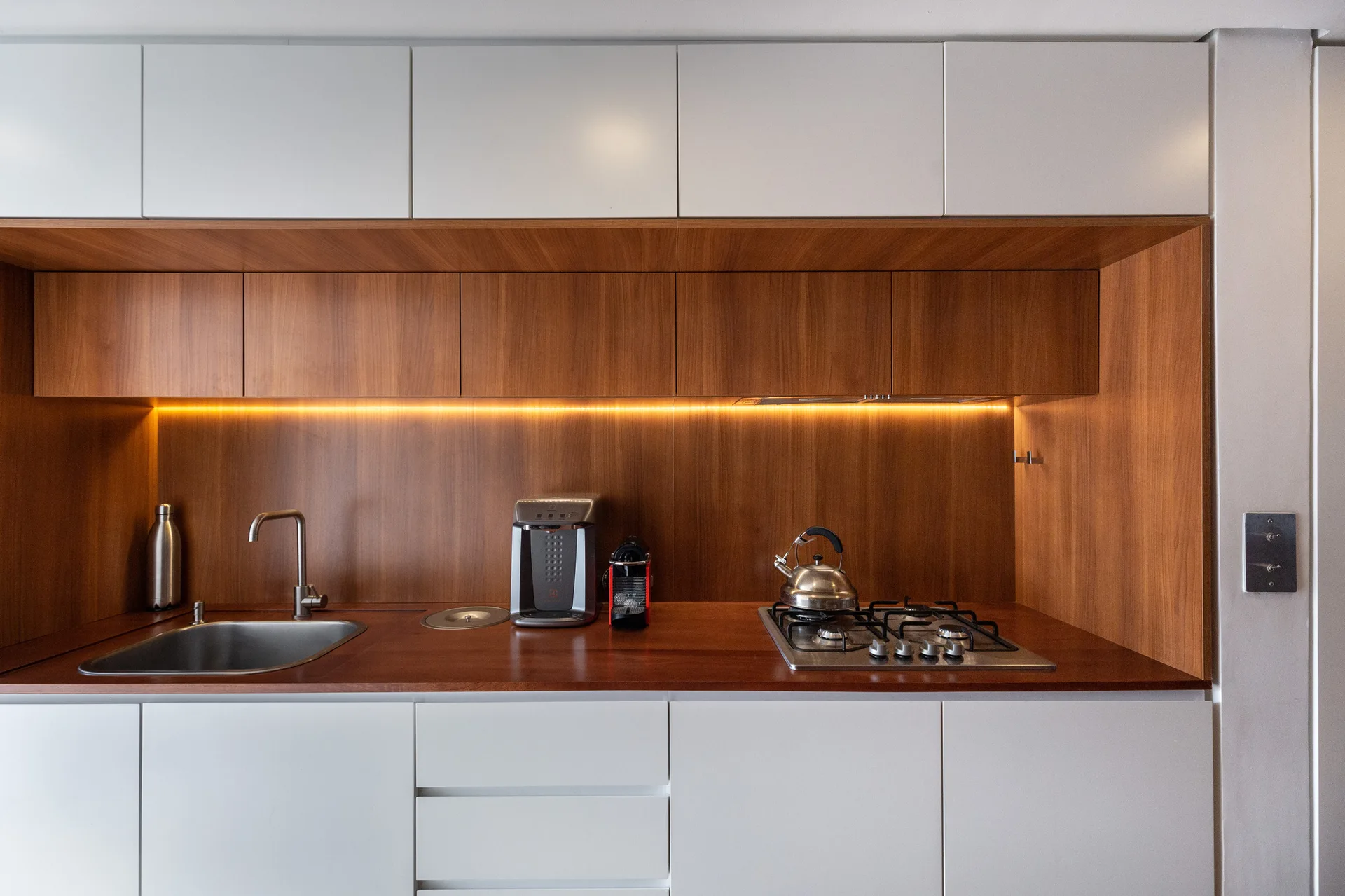 A minimalist kitchen features matte white cabinets flanking a warm wood-toned countertop and backsplash niche with integrated appliances and led lighting.