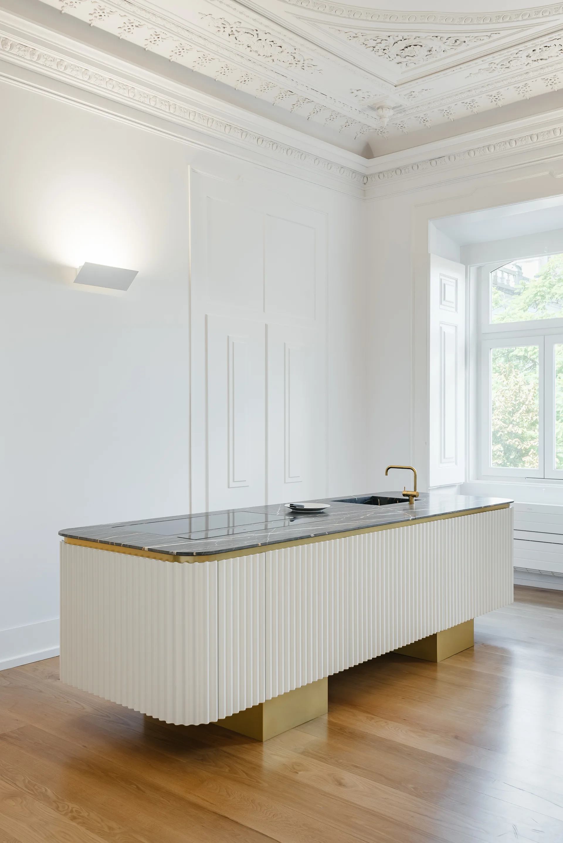 A minimalist kitchen island with a fluted off-white base and dark marble countertop stands on wood floors in a room with ornate molding.