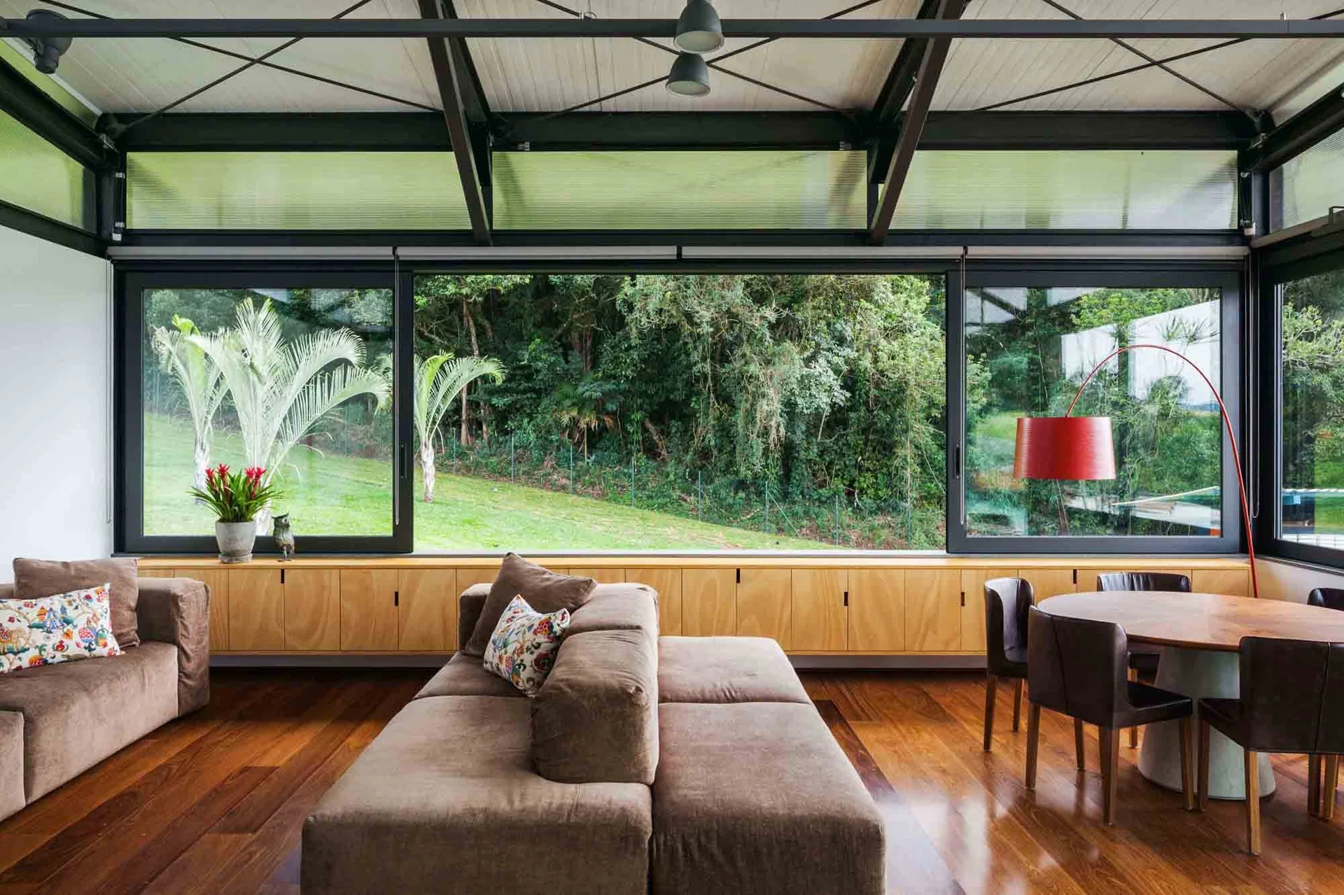 A modern living room with polished wood floors features a modular brown sofa, low wood cabinets under large glass windows, and a dining set.
