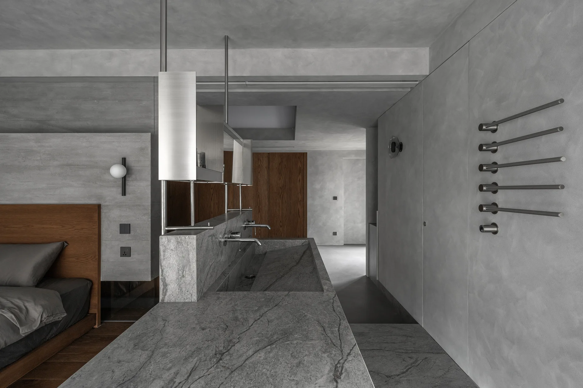A minimalist open-plan bedroom featuring a textured gray plaster wall, a wooden bed, and a long gray stone vanity with an integrated trough sink.
