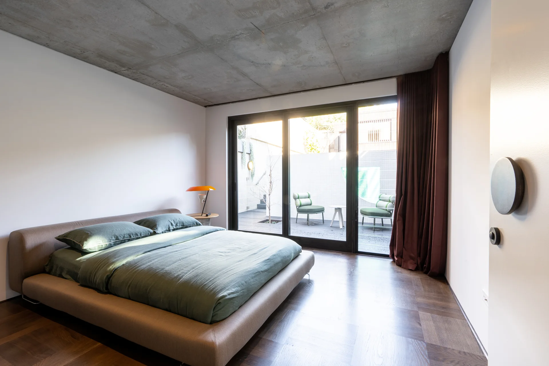 A modern bedroom features a low platform bed with green bedding under an exposed concrete ceiling next to sliding glass patio doors.