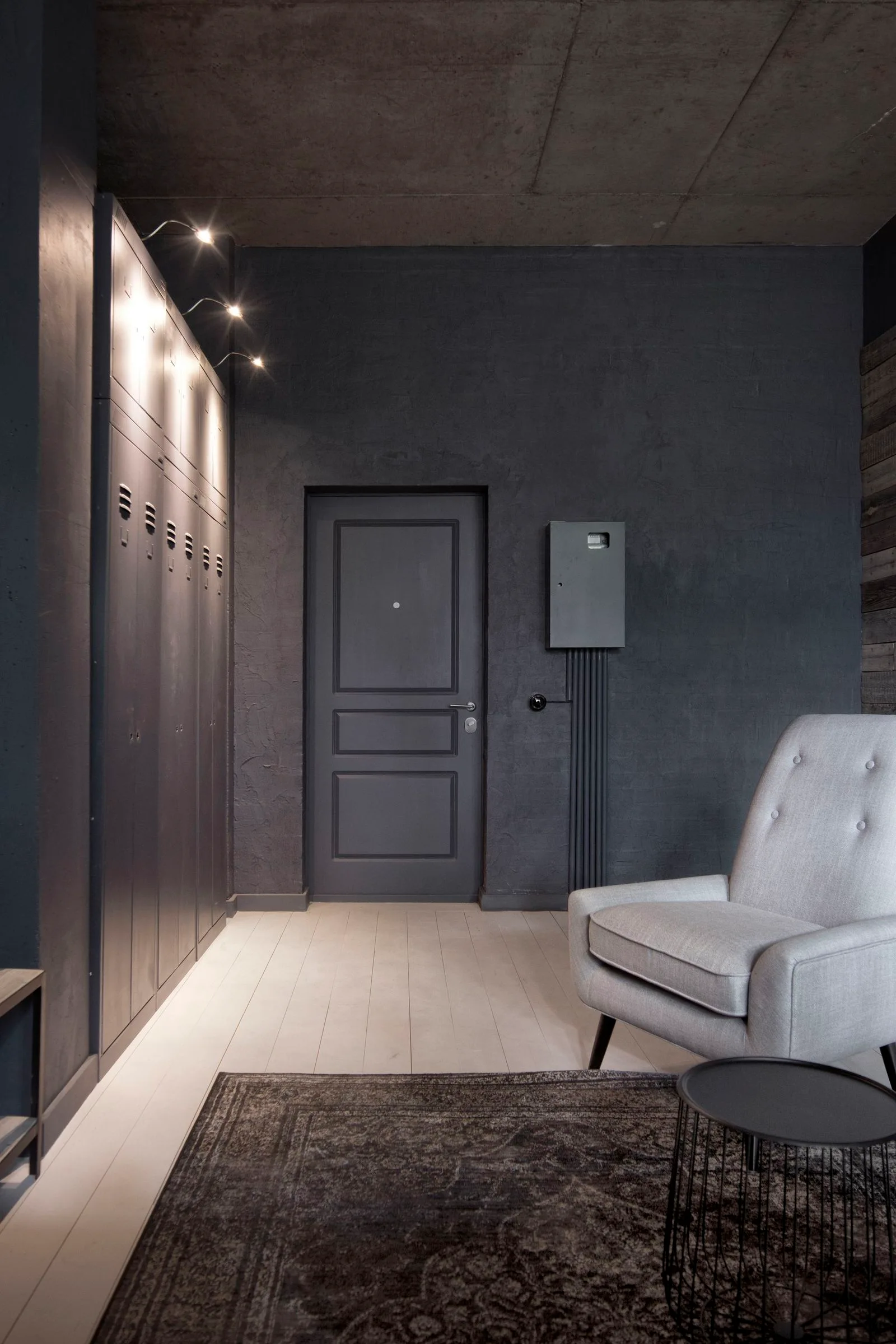 An industrial entryway with dark charcoal walls, a concrete ceiling, a bank of metal locker cabinets, a light wood floor, and a grey armchair.