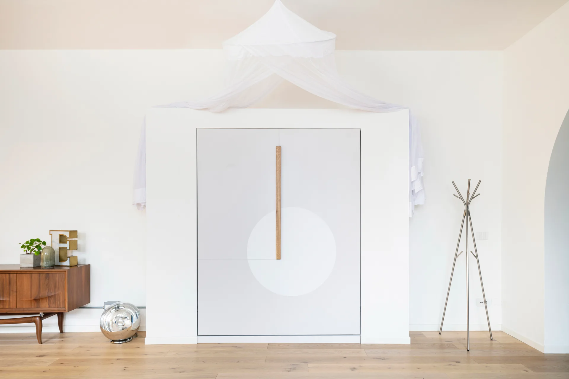 A minimalist white wardrobe with a fabric canopy sits on wood flooring next to a mid-century sideboard and a metallic coat rack.