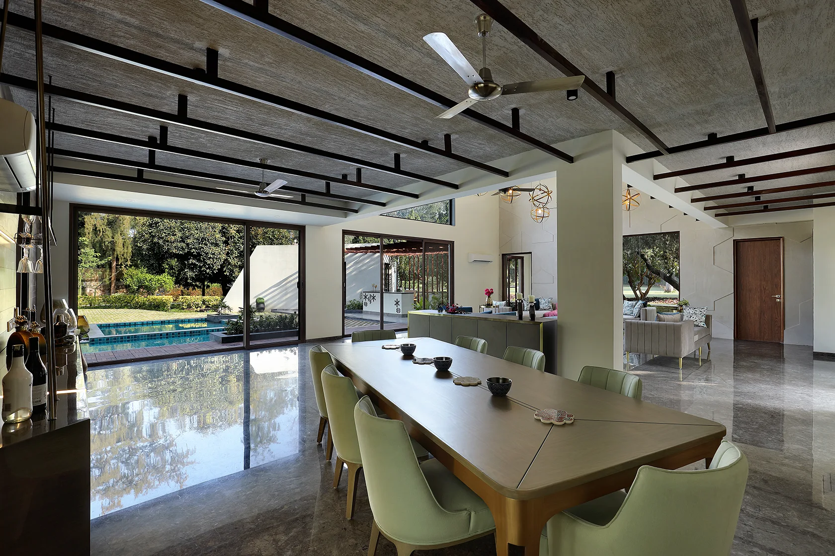 A spacious open dining room features a wood table with sage green chairs, a highly reflective stone floor, and dark ceiling beams.