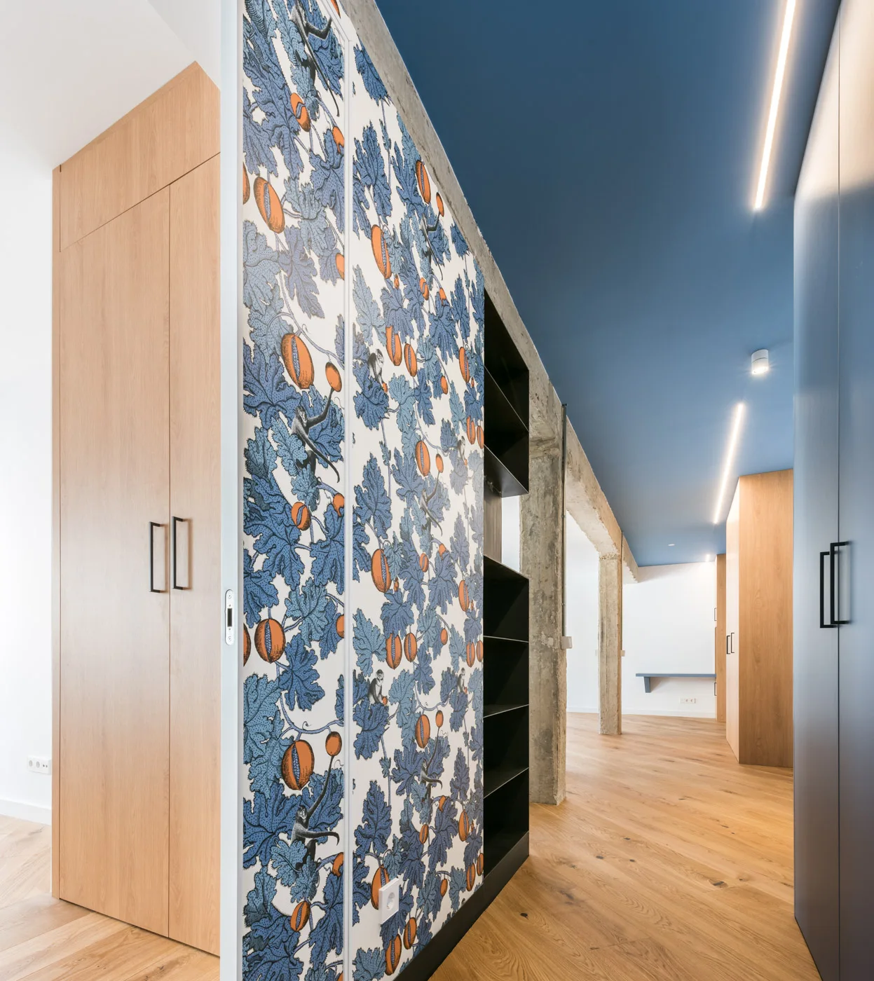 A modern hallway features light wood built-in cabinets, a section with blue botanical wallpaper, diagonal hardwood flooring, and a solid blue ceiling.