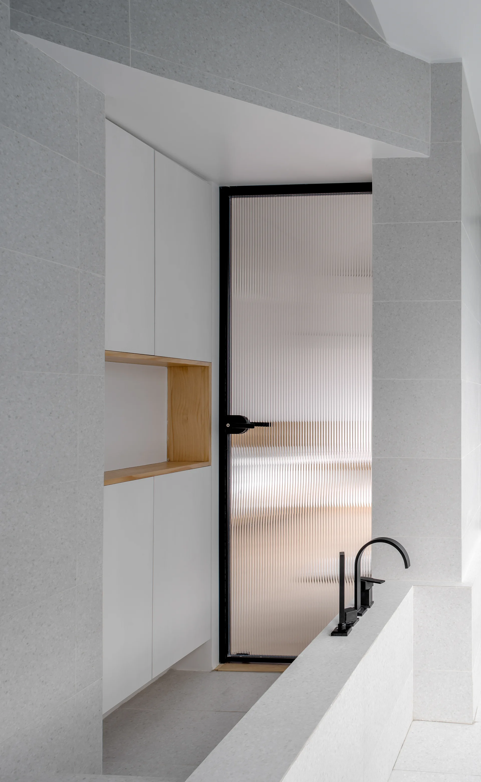 Modern bathroom with gray speckled tile walls, white built-in storage with a wooden niche, and a fluted glass door with black hardware.