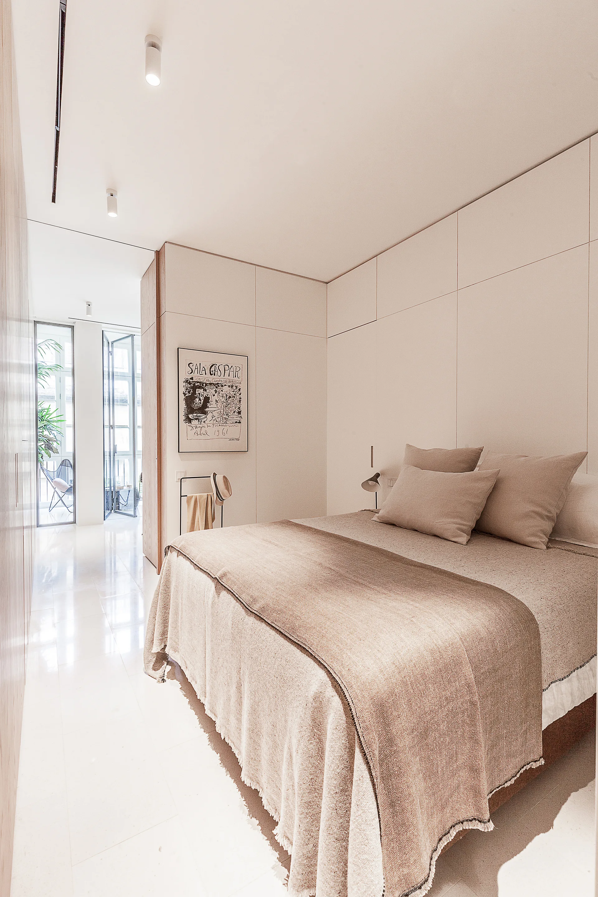 A minimalist bedroom features a bed dressed in neutral linen, integrated off-white wall panels, and glossy floors leading to a glass patio door.