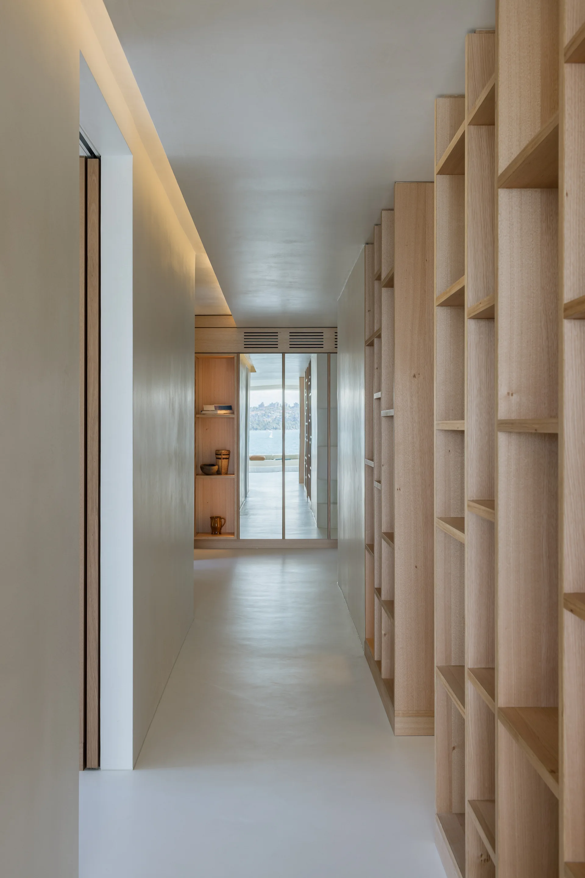 A minimalist hallway features light wood built-in shelving along one wall and a mirrored door at the end reflecting a coastal water view.