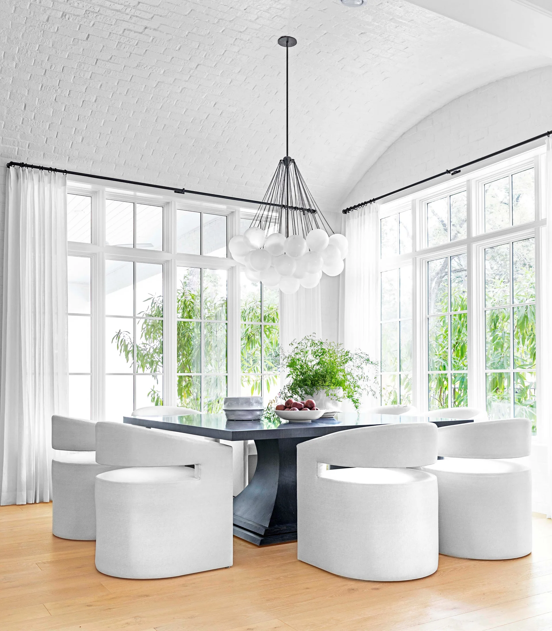 A bright dining room featuring a dark square dining table and white barrel chairs under a white vaulted brick ceiling and a bubble chandelier.