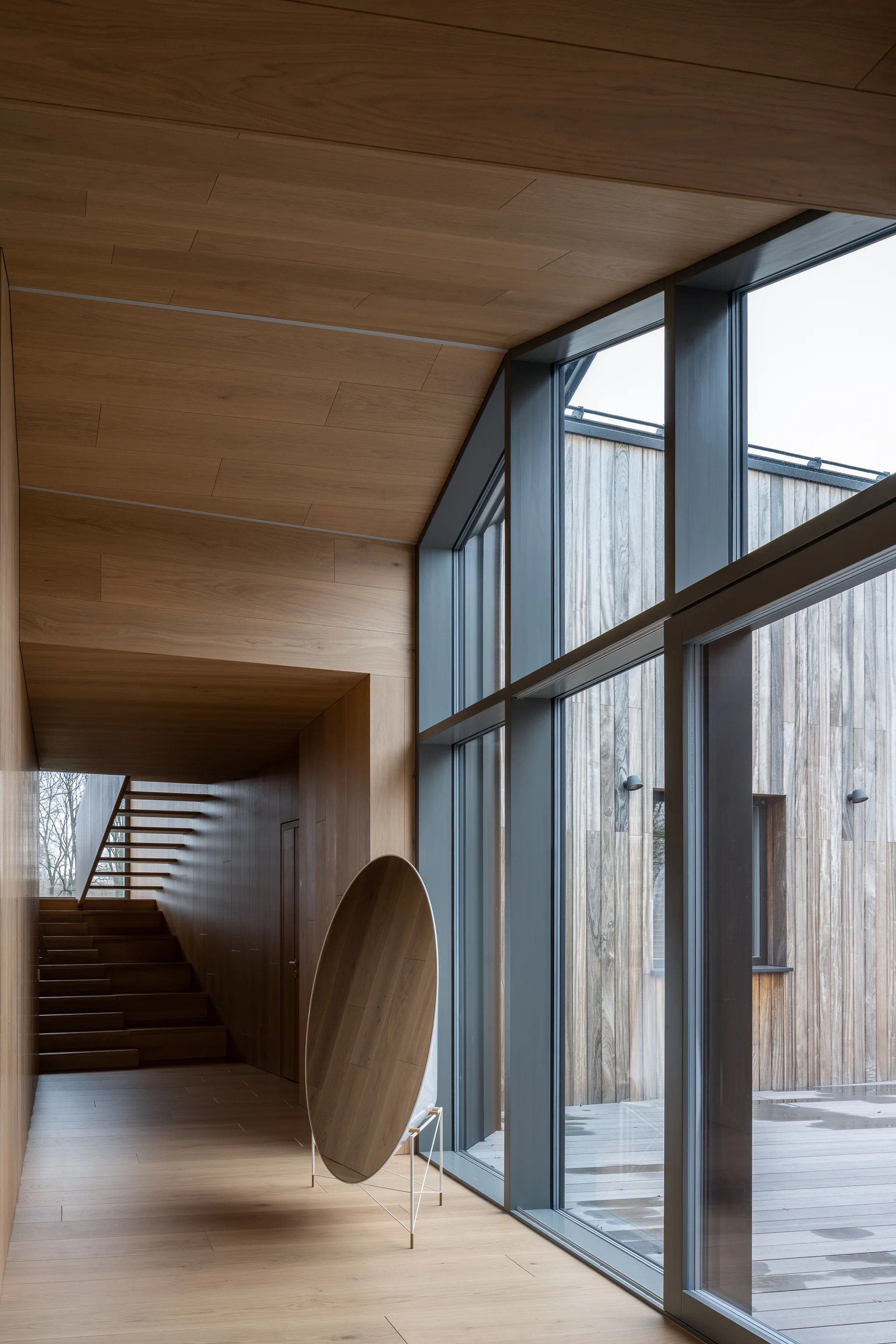 A contemporary hallway with seamless light wood wall paneling, a wooden staircase, and a large metal-framed glass window overlooking a deck.