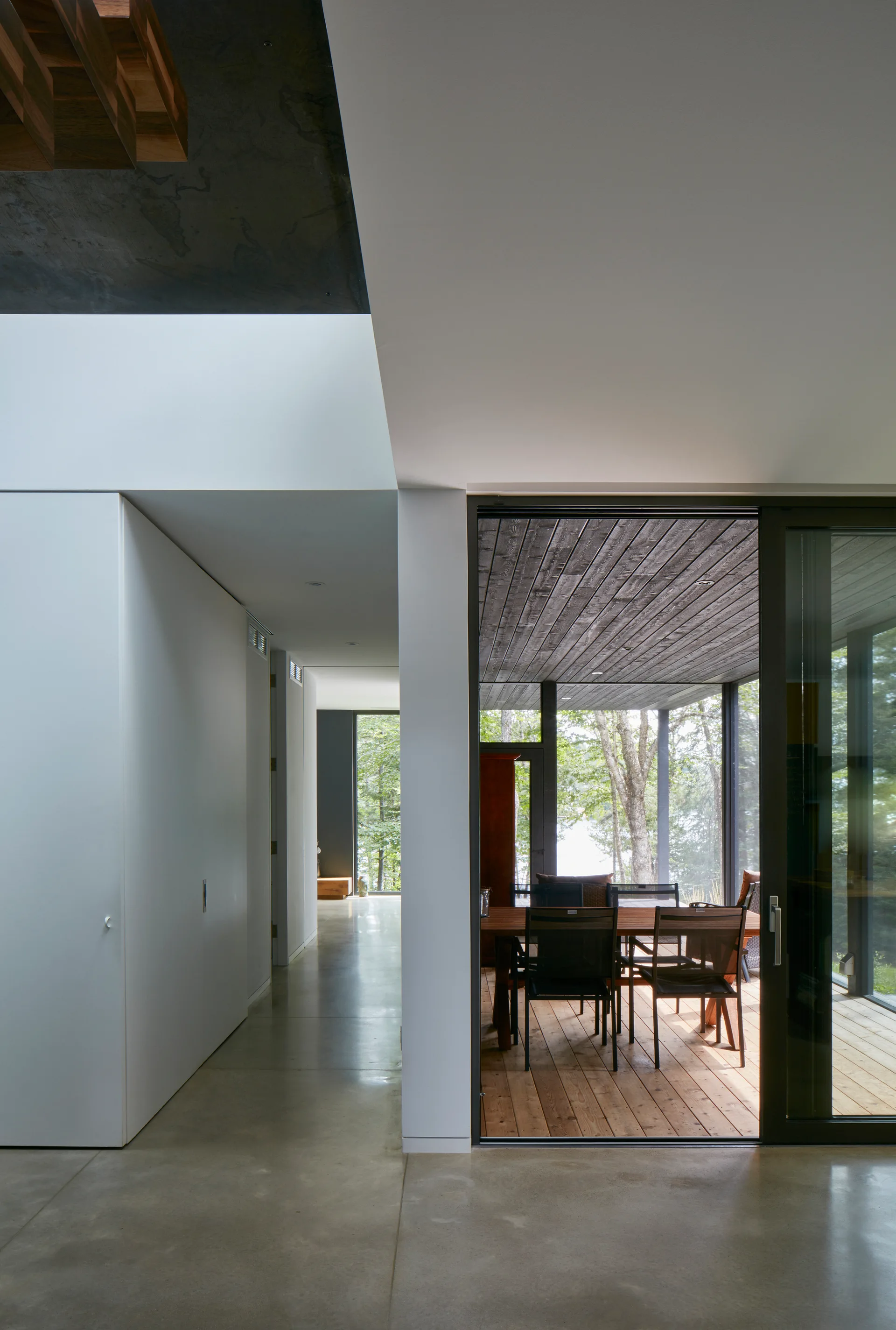 A minimalist hallway with polished concrete flooring and white walls leads to a sliding glass door opening onto a wooden dining deck.