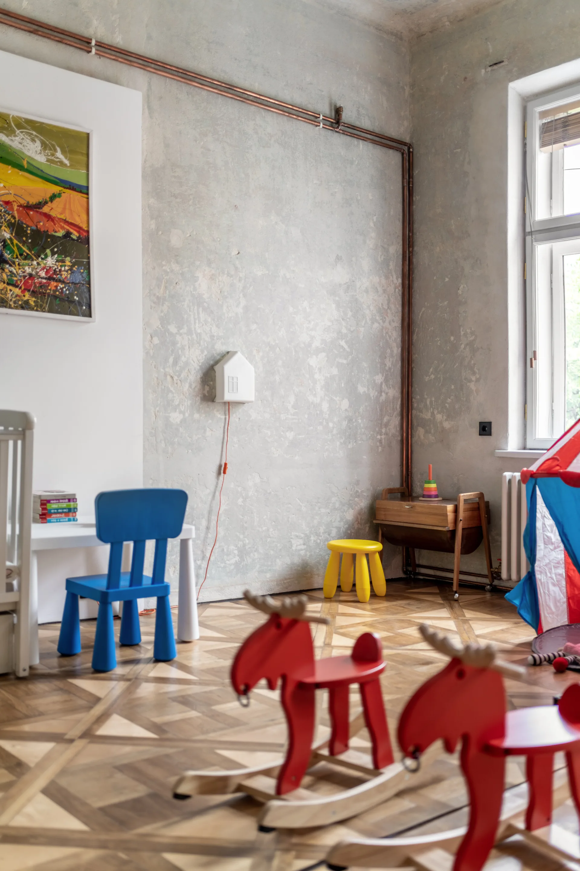 A kids playroom features raw grey plaster walls, exposed copper piping, patterned parquet wood flooring, and colorful child-sized furniture.
