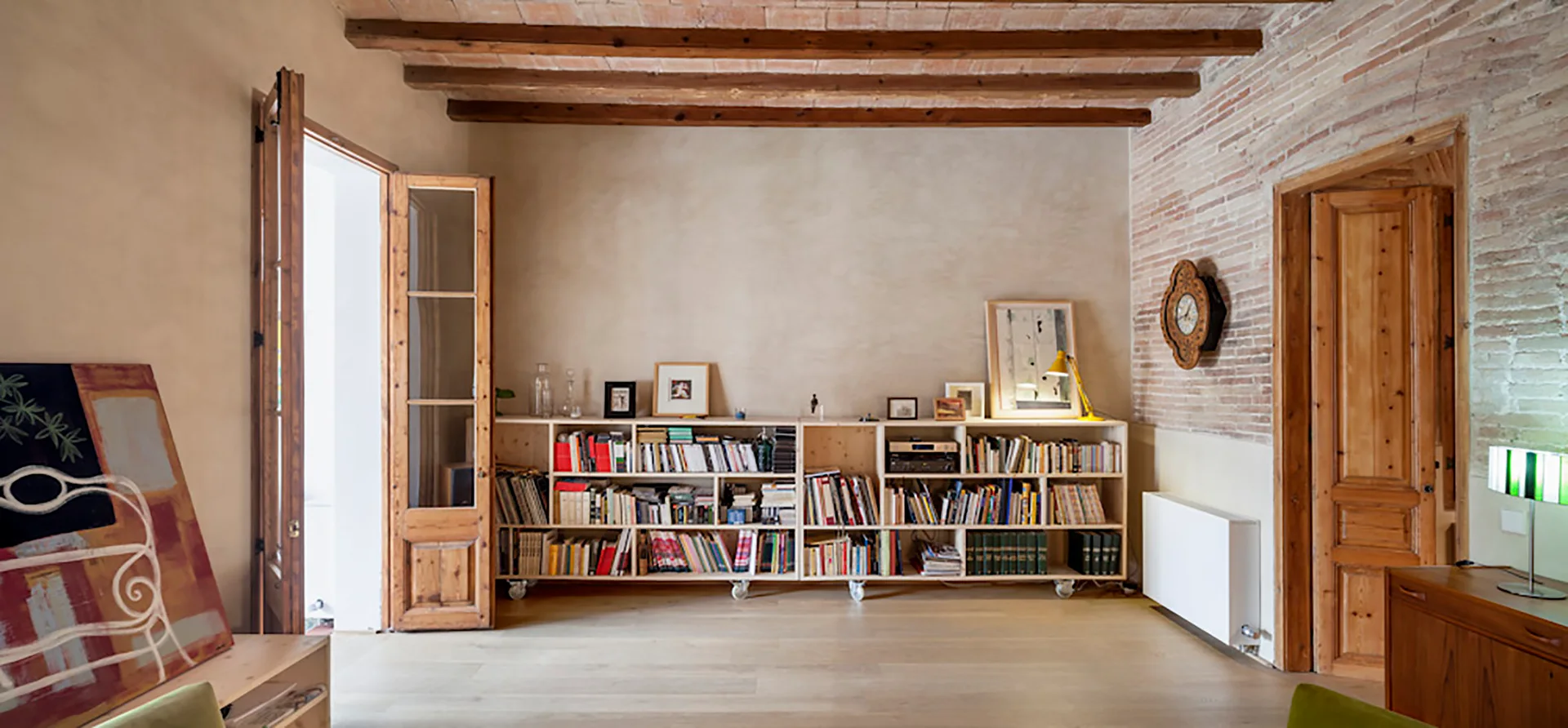 A rustic living room featuring a low wooden bookcase on wheels, exposed ceiling beams, a plaster wall, and a brick accent wall.