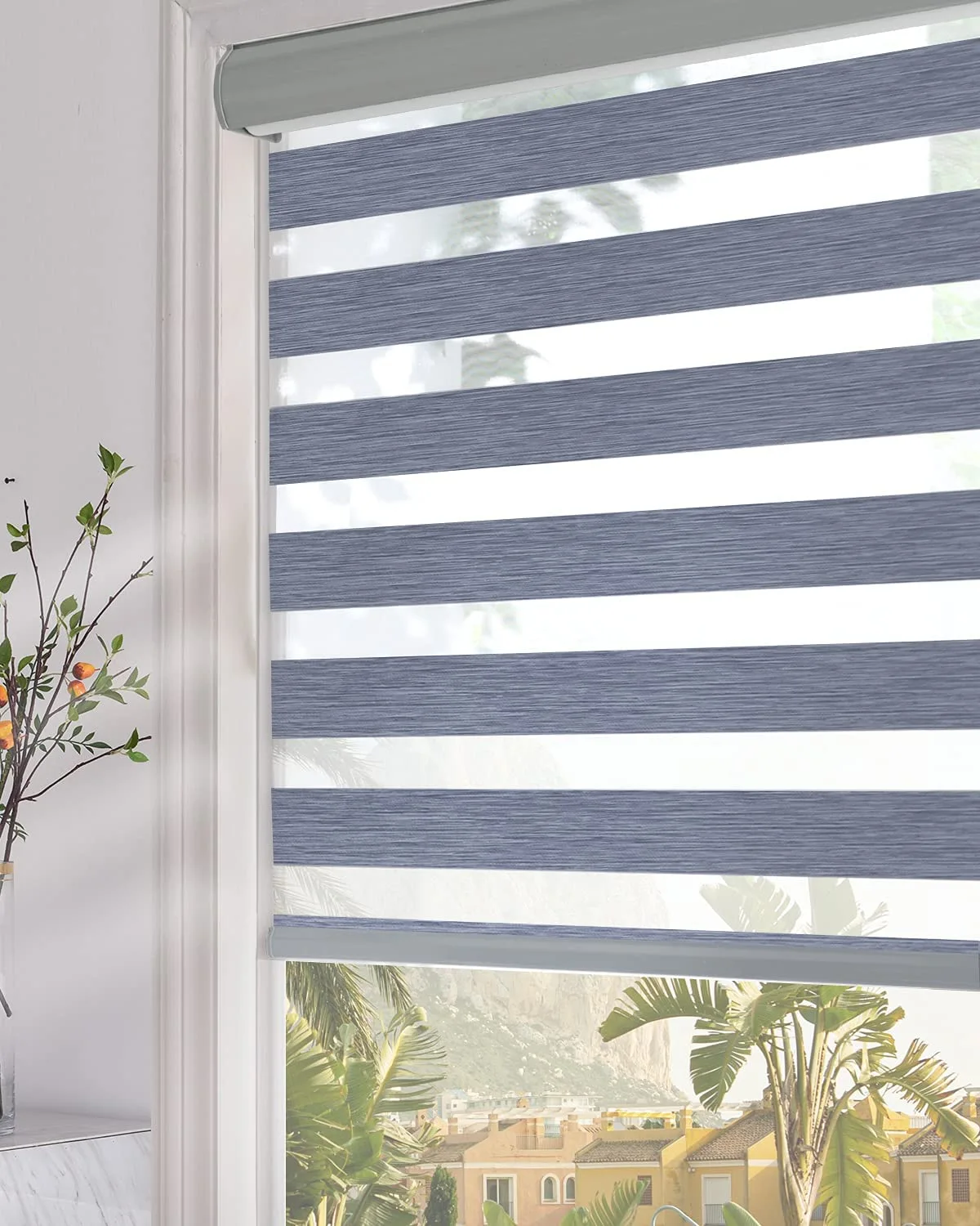 A close-up of a blue and white striped zebra roller blind installed on a white window frame with a leafy green branch in a glass vase nearby.