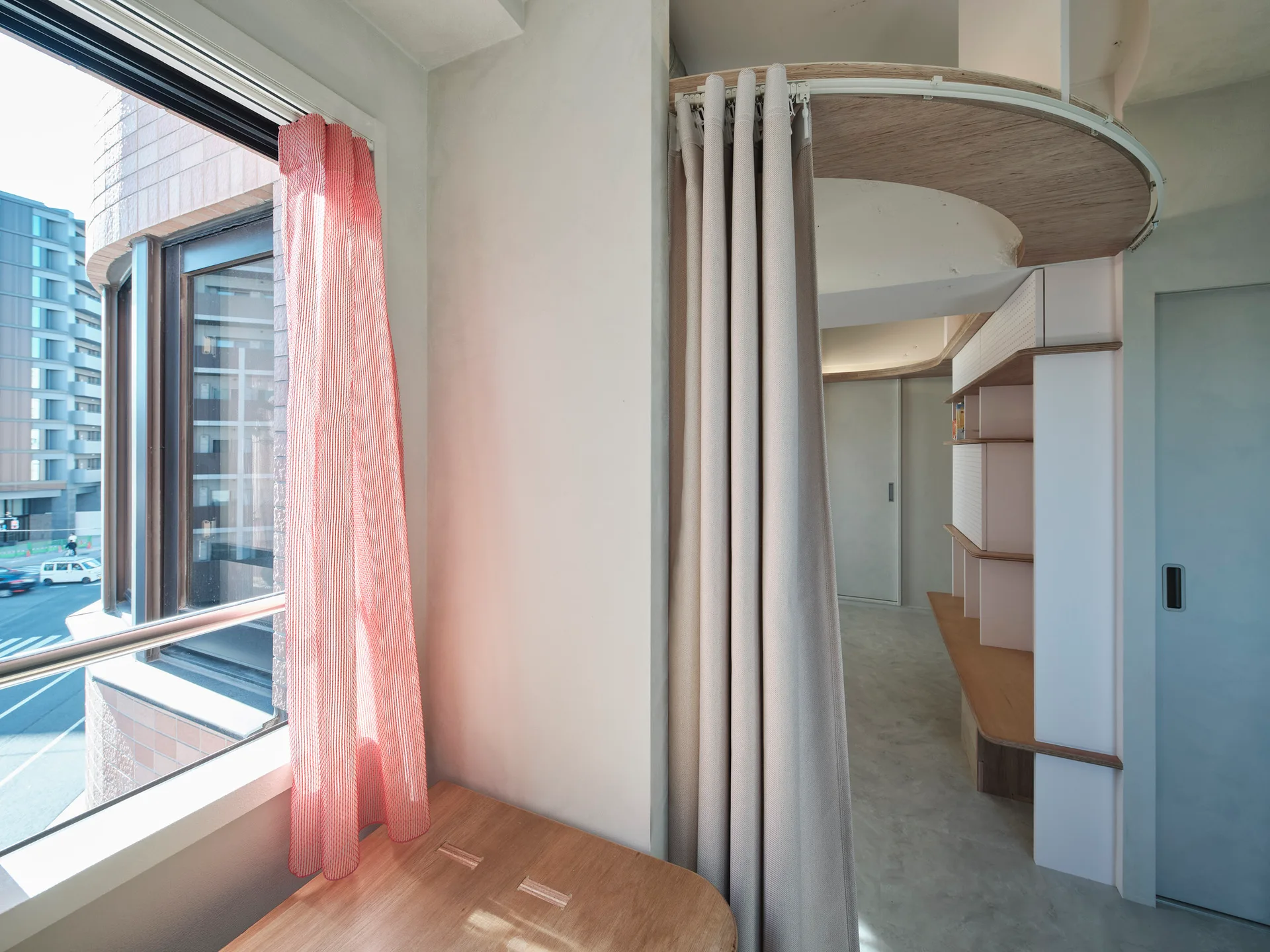 A minimalist room features custom light wood shelving and a curved ceiling track with a beige curtain next to a window with a red curtain.