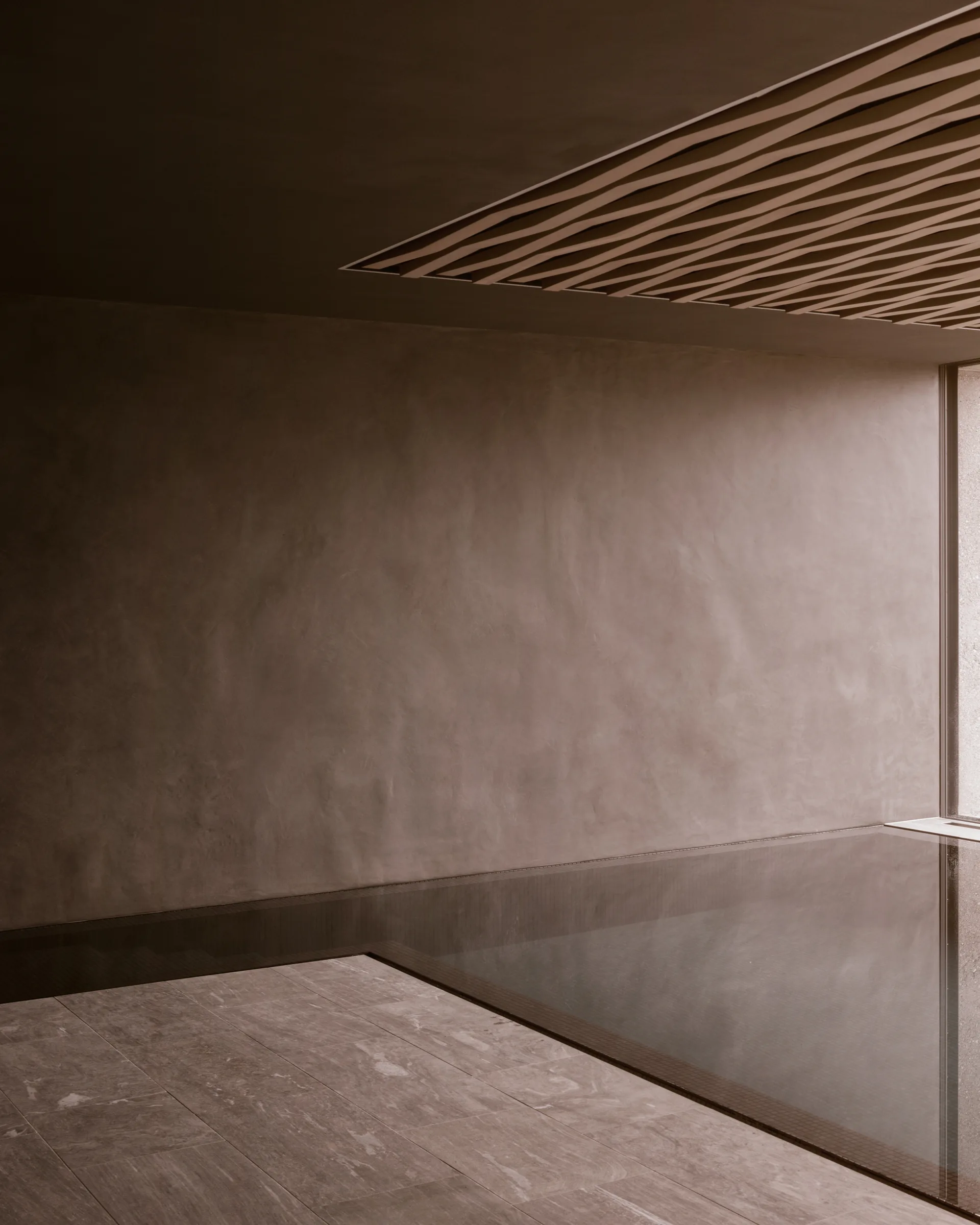 A minimalist indoor pool area with textured brown plaster walls, stone tile flooring, and a decorative slatted wooden ceiling panel.