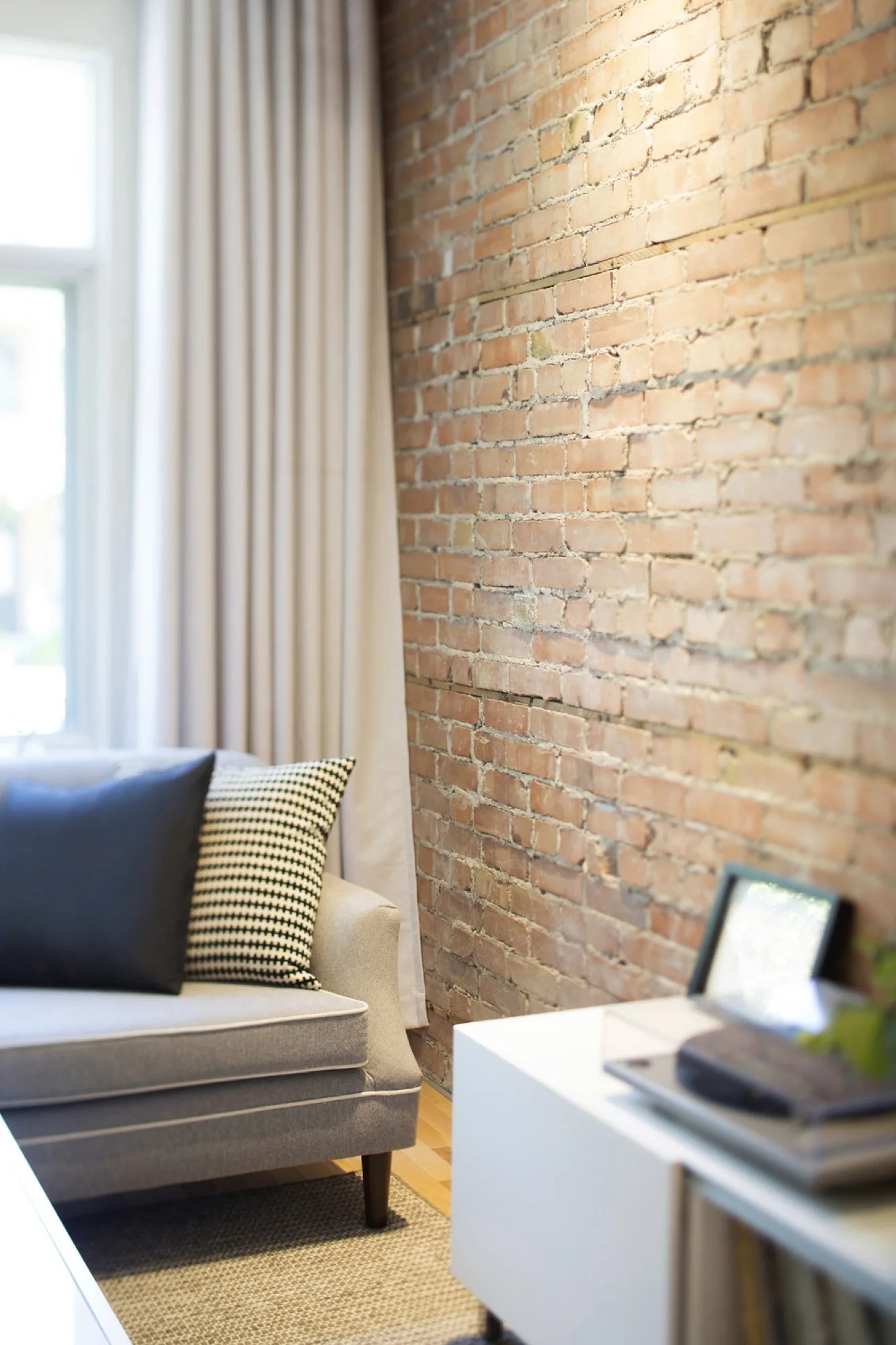 A grey upholstered armchair with decorative pillows sits beside an exposed brick wall and long beige curtains in a sunlit room.