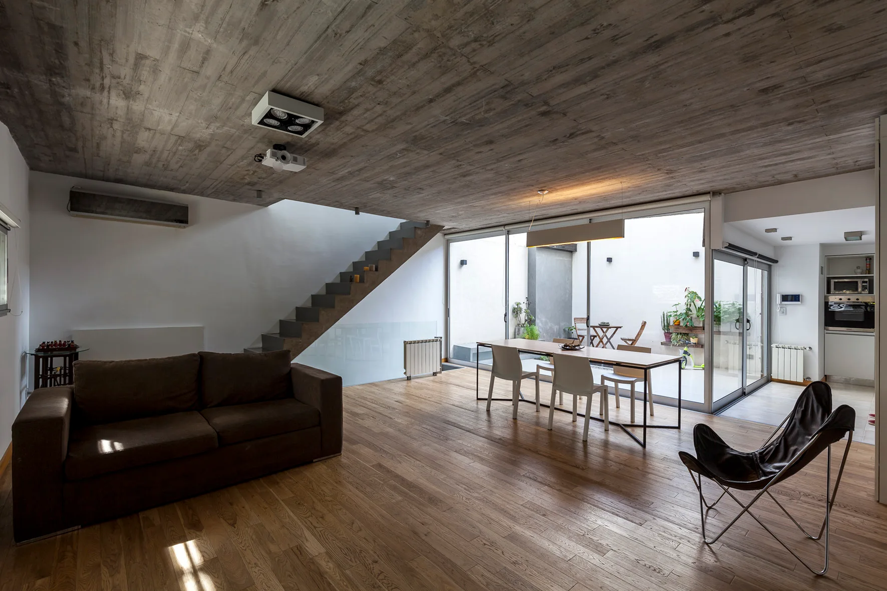 An open-plan living and dining area featuring a textured concrete ceiling, wooden flooring, a brown sofa, and sliding glass doors opening to a courtyard.