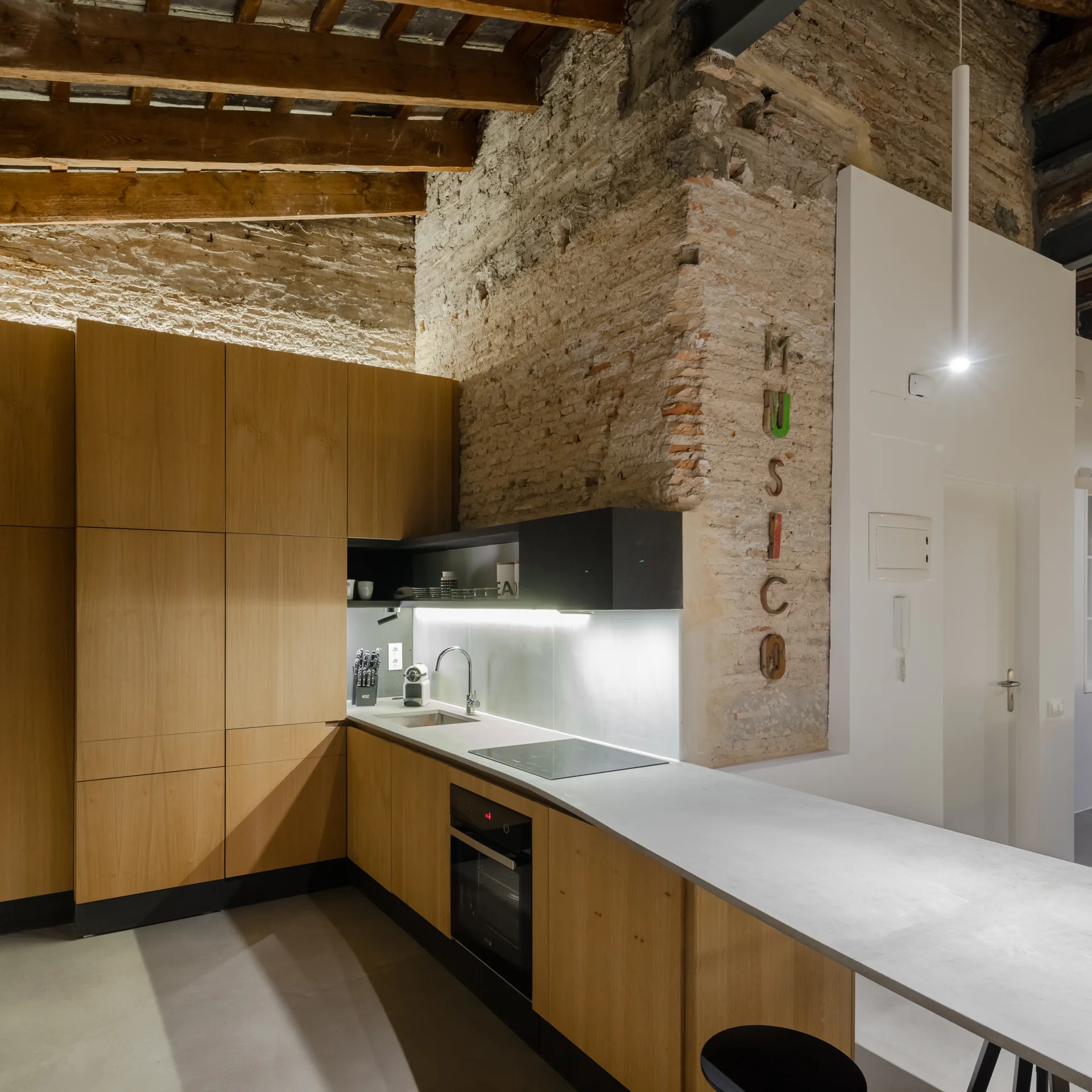 A modern kitchen featuring flat-front wood cabinets, a light gray countertop, and an exposed brick accent wall under rustic wood ceiling beams.
