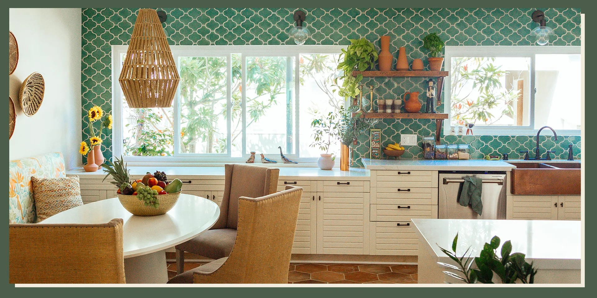 A bright bohemian kitchen featuring green patterned wall tiles, white slatted cabinetry under large windows, and a woven pendant light over a dining table.