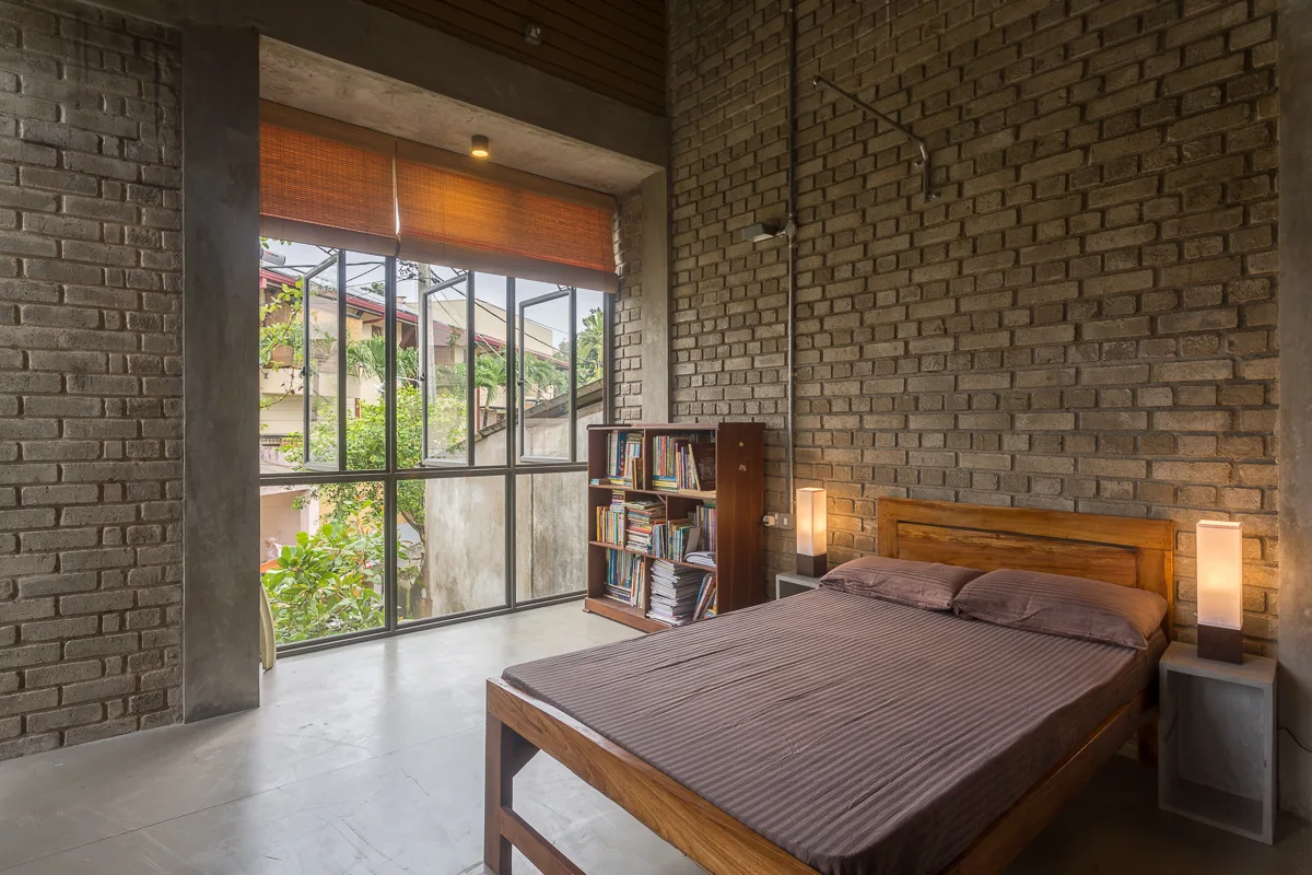 An industrial bedroom featuring a wooden bed, gray exposed brick walls, a large metal-framed window with woven blinds, and a bookcase.