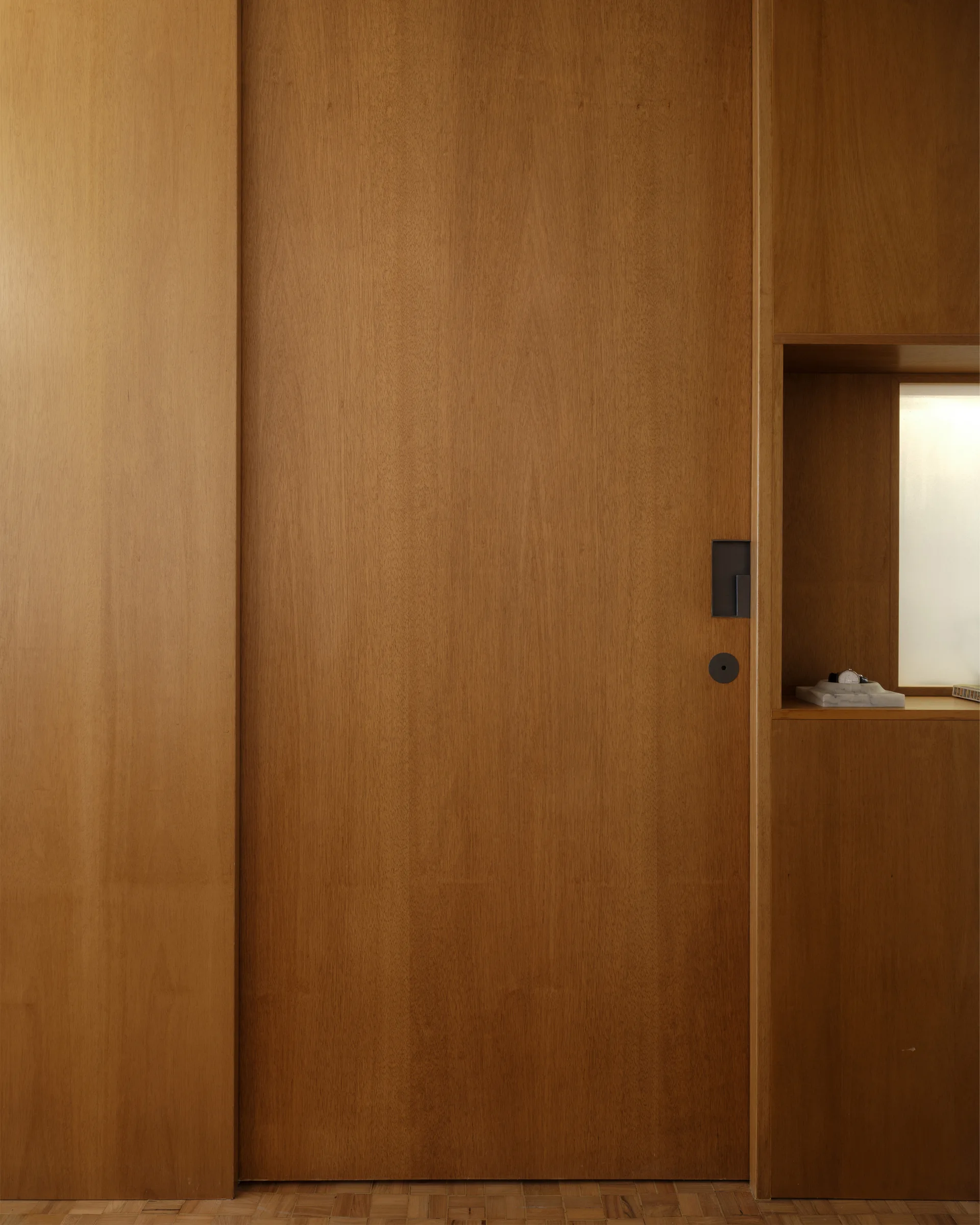 A seamless wood-paneled wall includes a flush door with a dark rectangular handle and a small lighted niche holding a decorative bowl.
