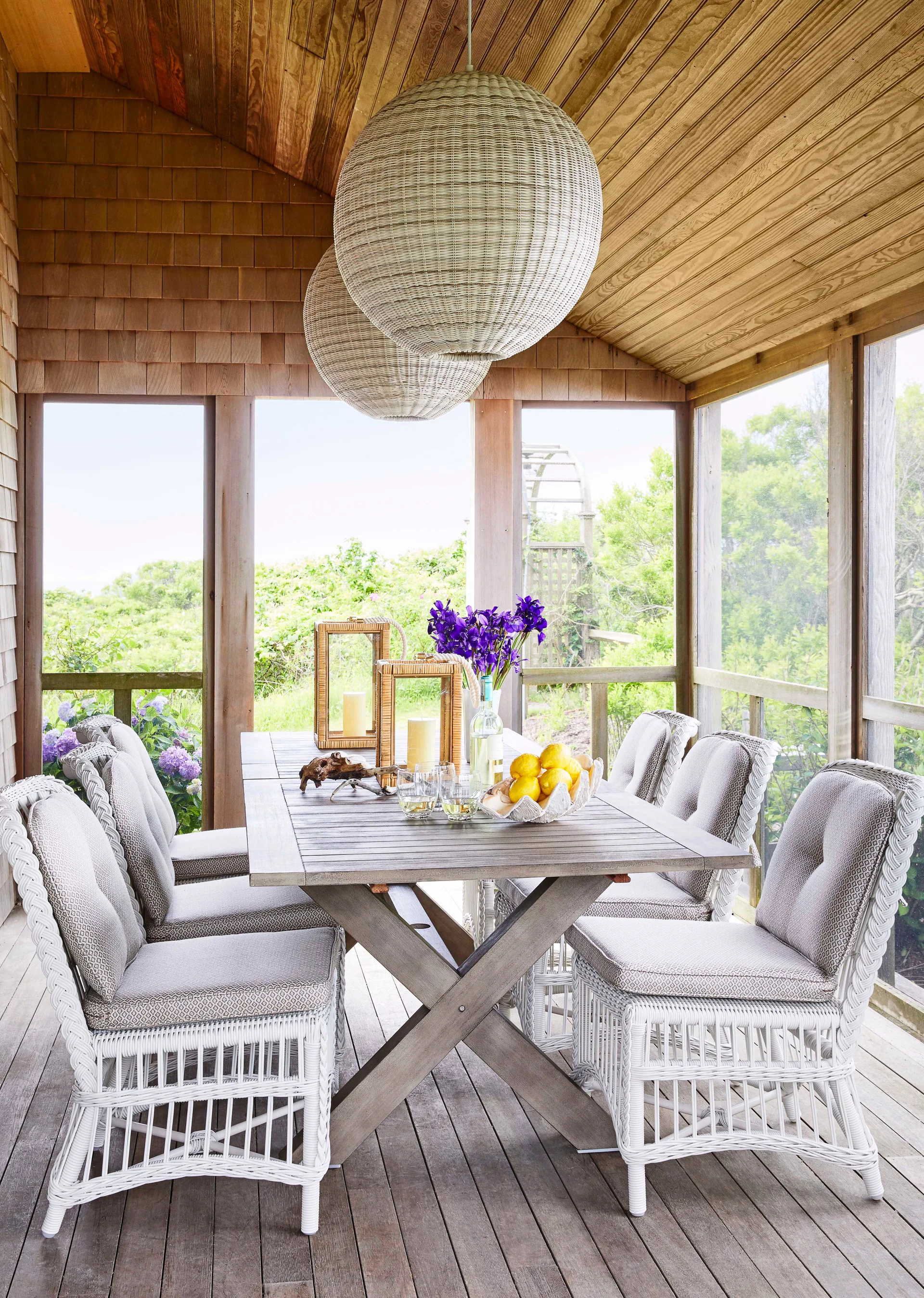A wooden dining table with white wicker chairs on a screened porch under two large woven spherical pendant lights.