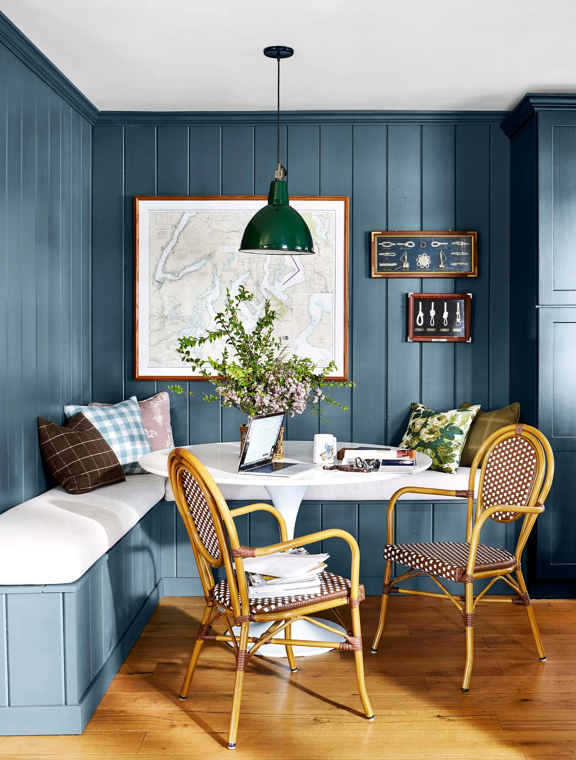 A breakfast nook with dark blue wood-paneled walls features a built-in banquette, a white pedestal table, and two woven rattan bistro chairs.