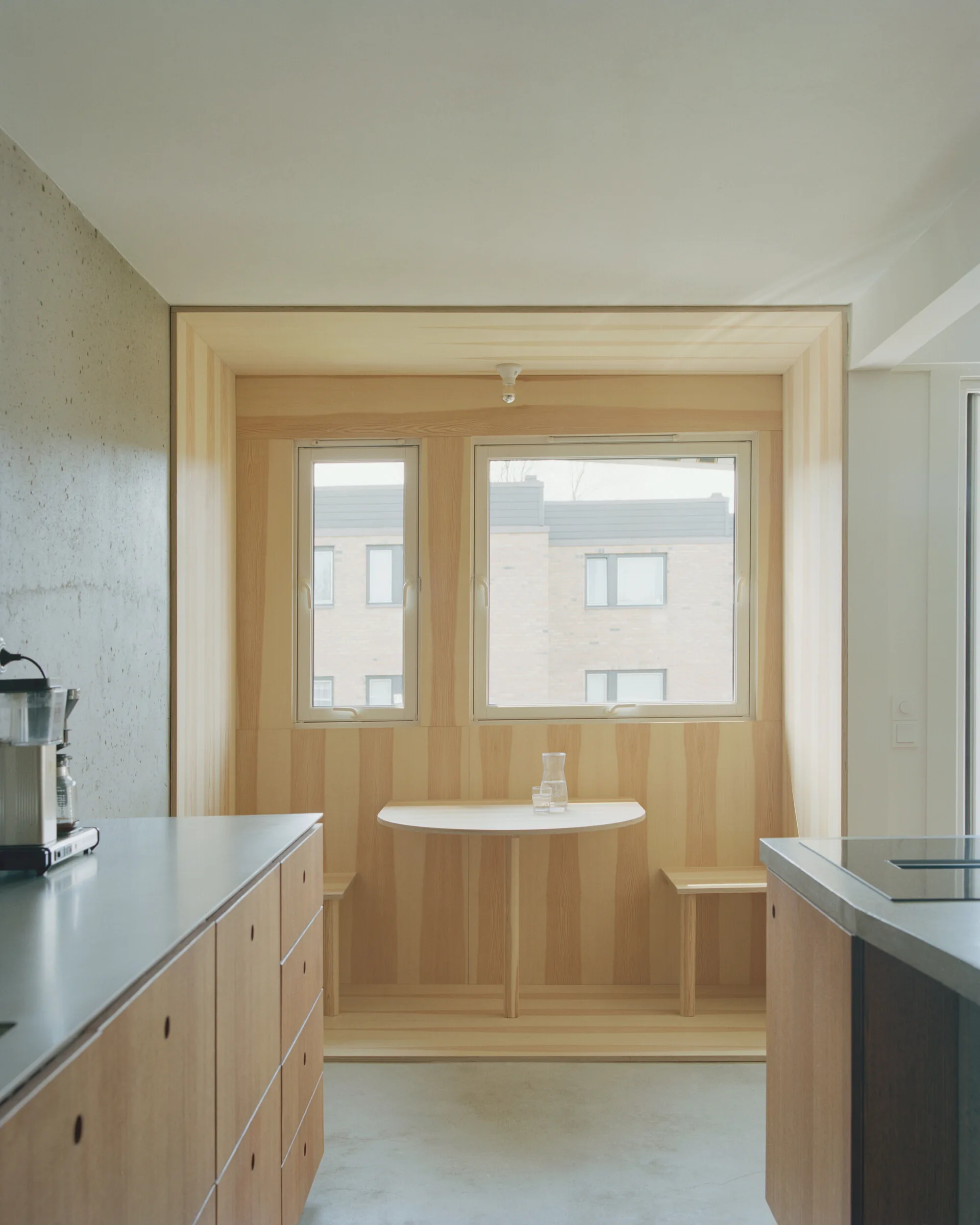 A modern kitchen with wood cabinetry, concrete flooring, and a recessed dining nook lined with vertical wood paneling around two windows.