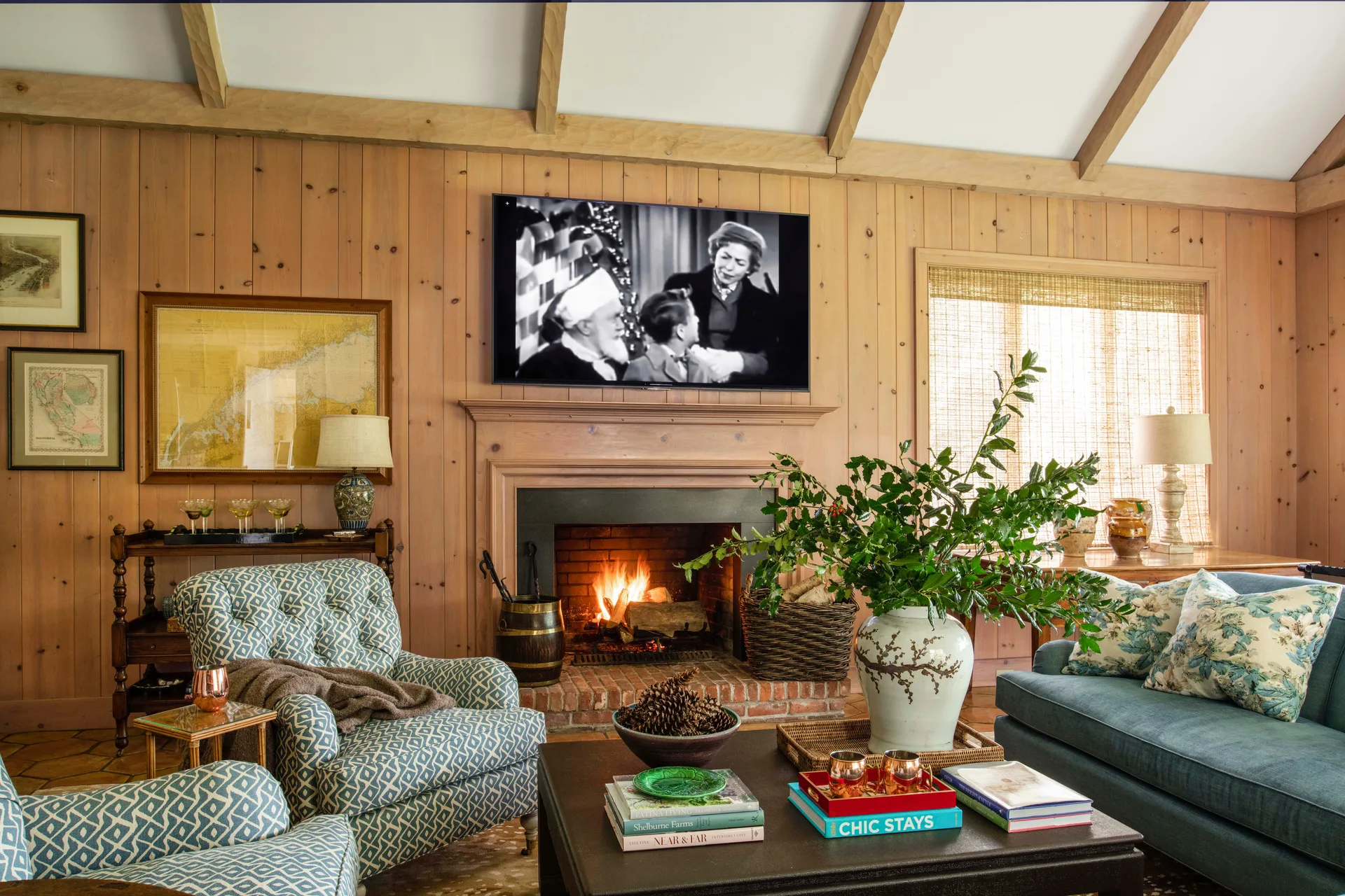 A rustic living room with vertical wood paneling, a burning brick fireplace, blue upholstered seating, and a vaulted ceiling with beams.