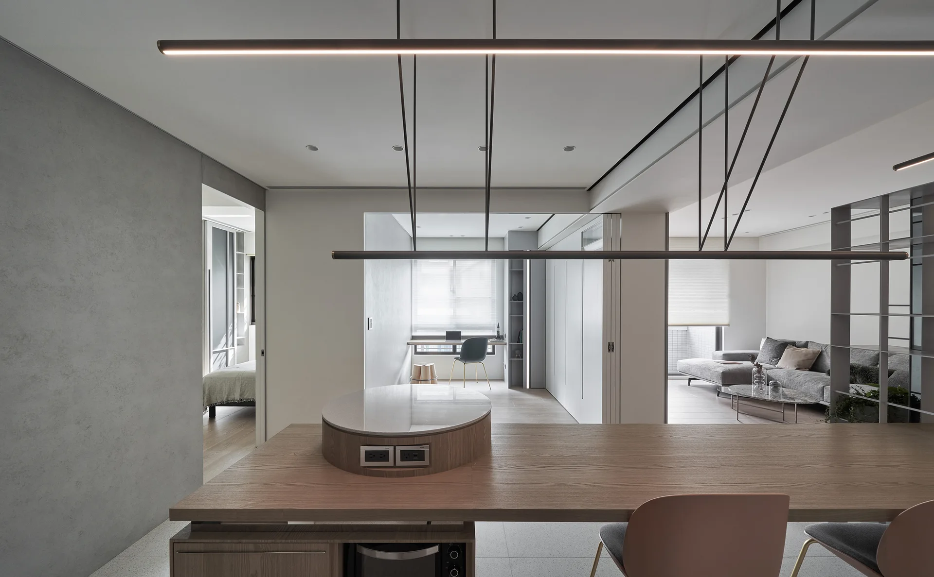 A modern open-plan dining space with a wooden table featuring a stone outlet hub, suspended linear light fixtures, and a view into an adjacent home office.