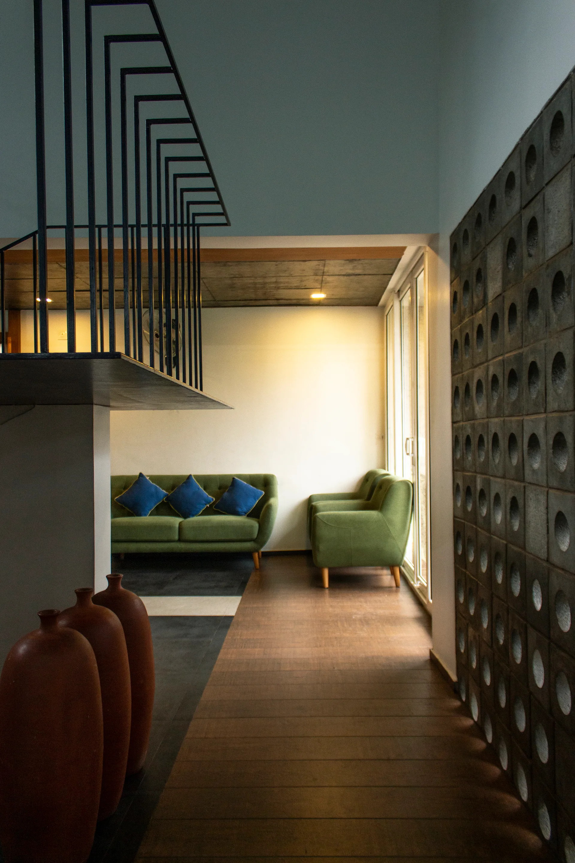 A modern living space features green sofas, a dark concrete screen wall with circular cutouts, and three tall terracotta vases on the floor.