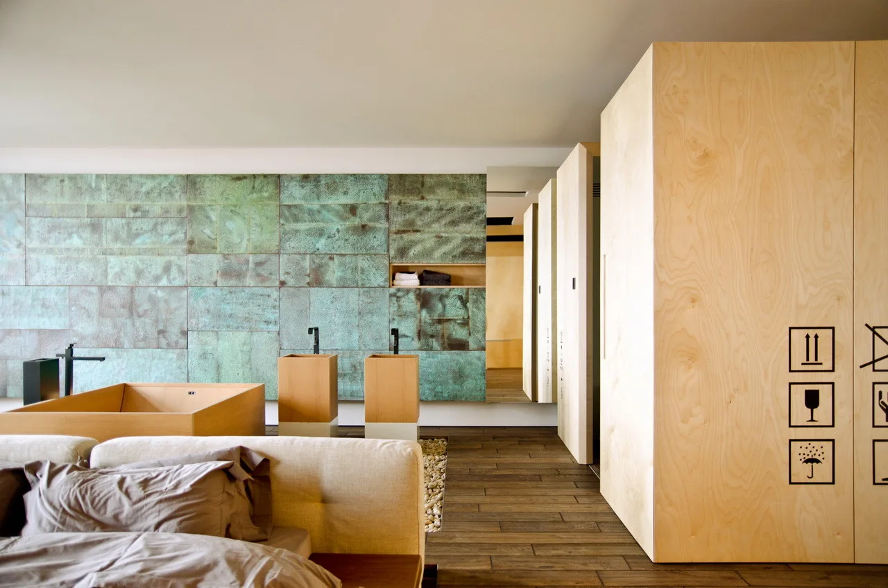 A modern open bedroom features a wooden bathtub and dual pedestal sinks set against an green oxidized copper tile wall next to large plywood cabinets.