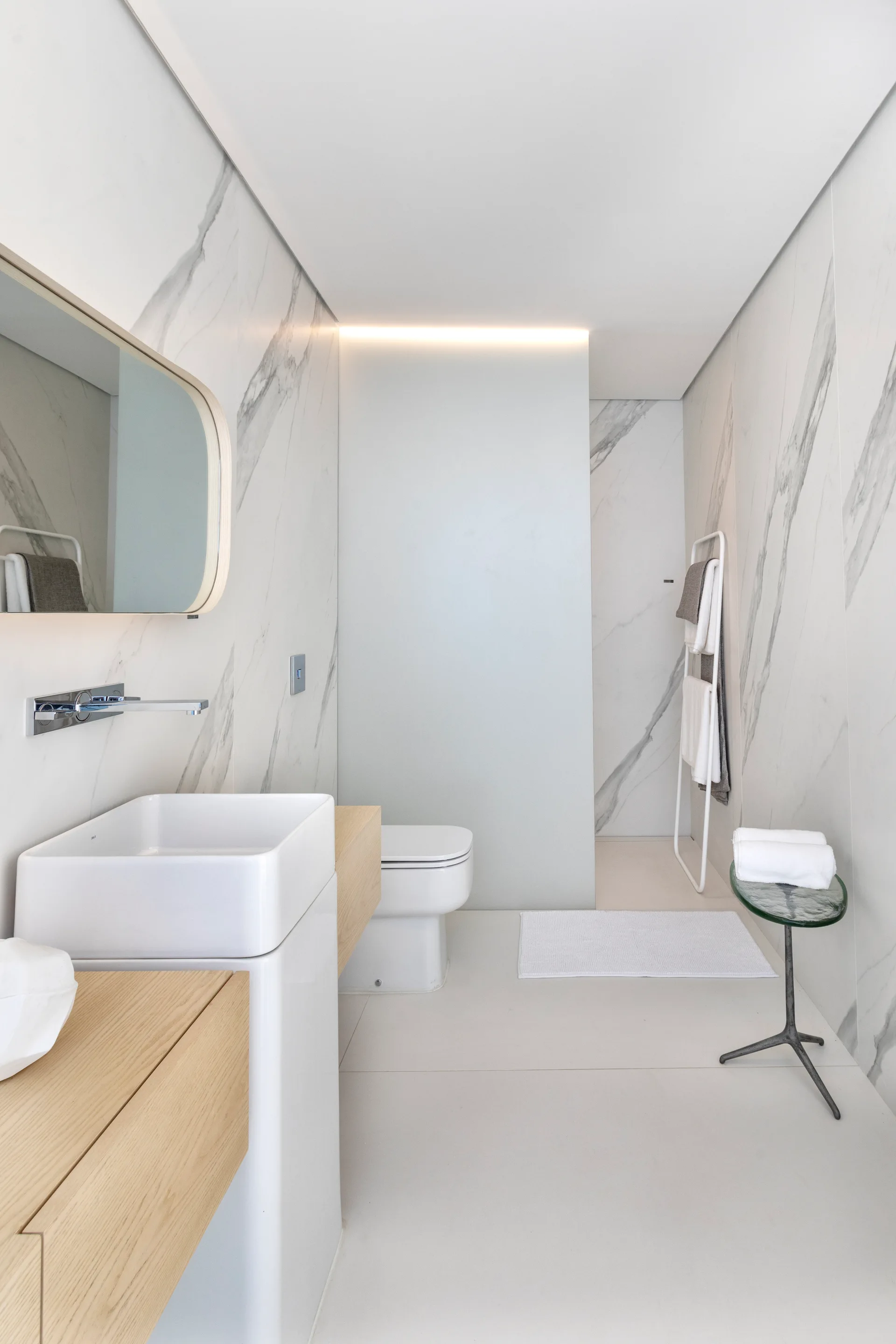A minimalist bathroom featuring marble-veined wall tiles, a white vessel sink on a light wood floating vanity, and a ladder towel rack.