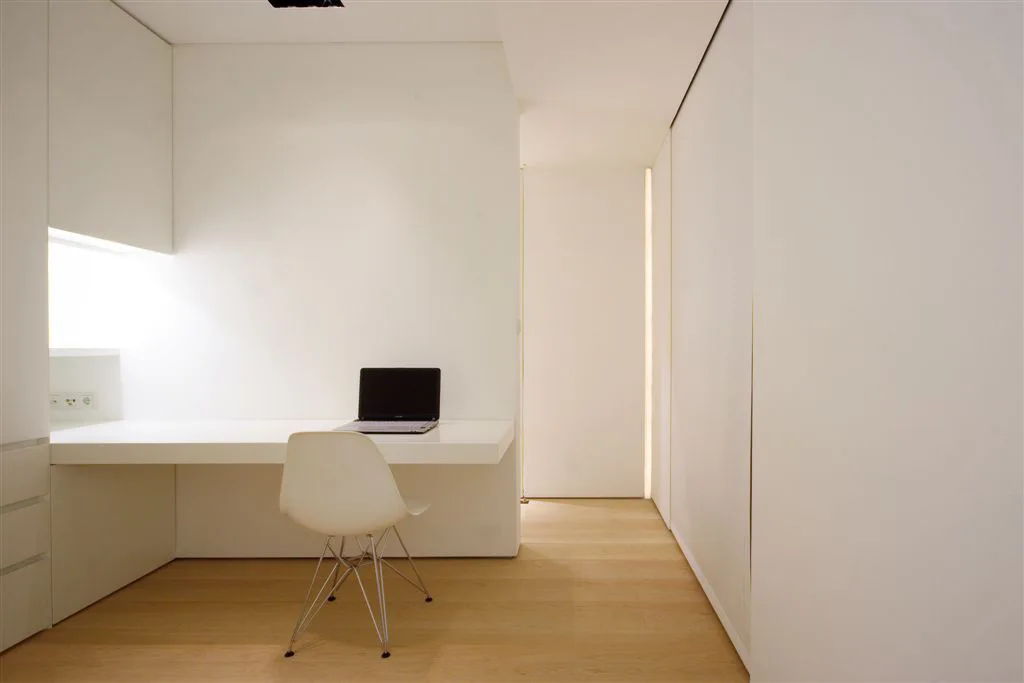 A minimalist home office features a white built-in desk with a laptop, a white molded plastic chair, and light wood flooring.