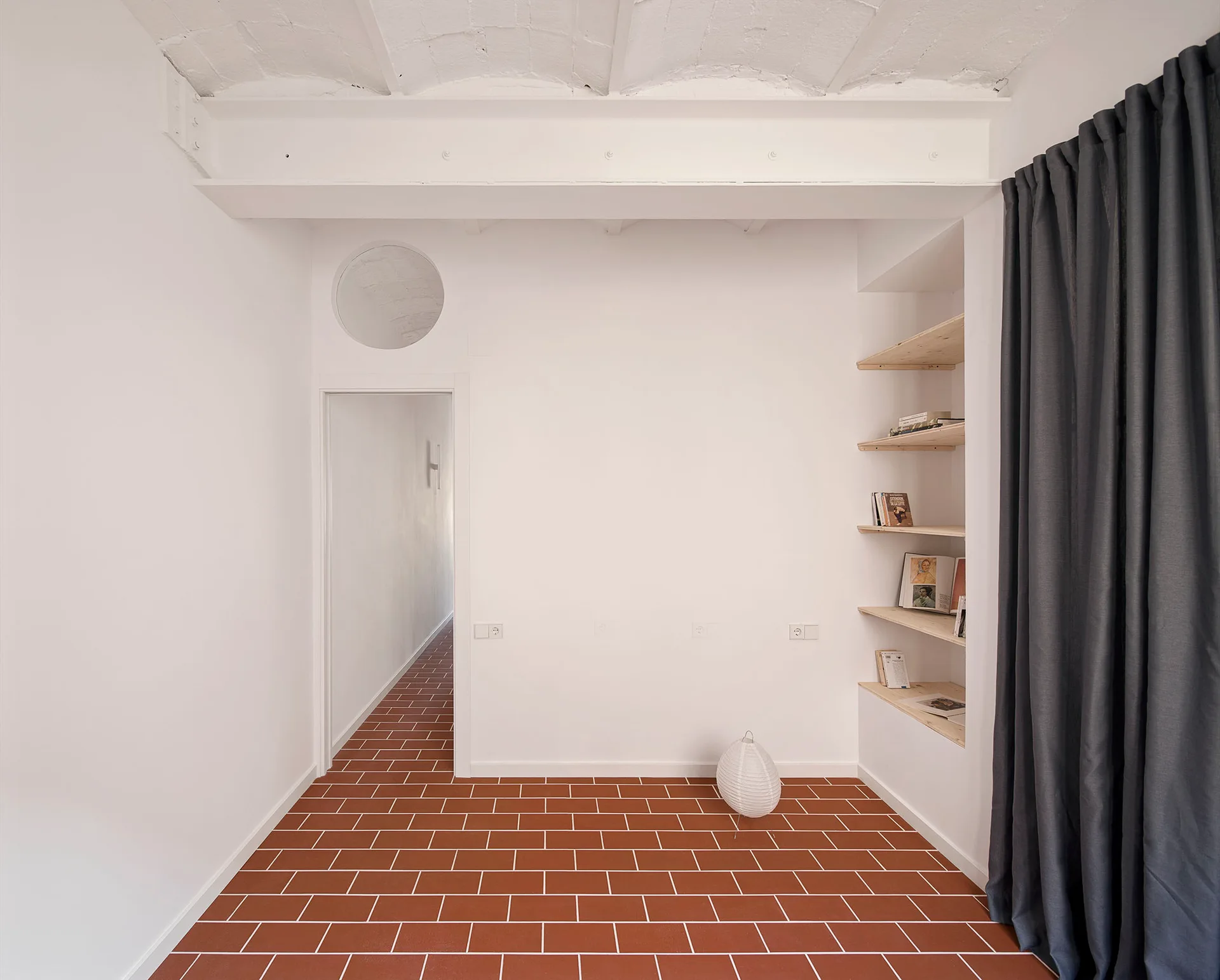 Minimalist interior featuring red terracotta floor tiles, a white vaulted ceiling, built-in wooden shelves, and a dark grey curtain.