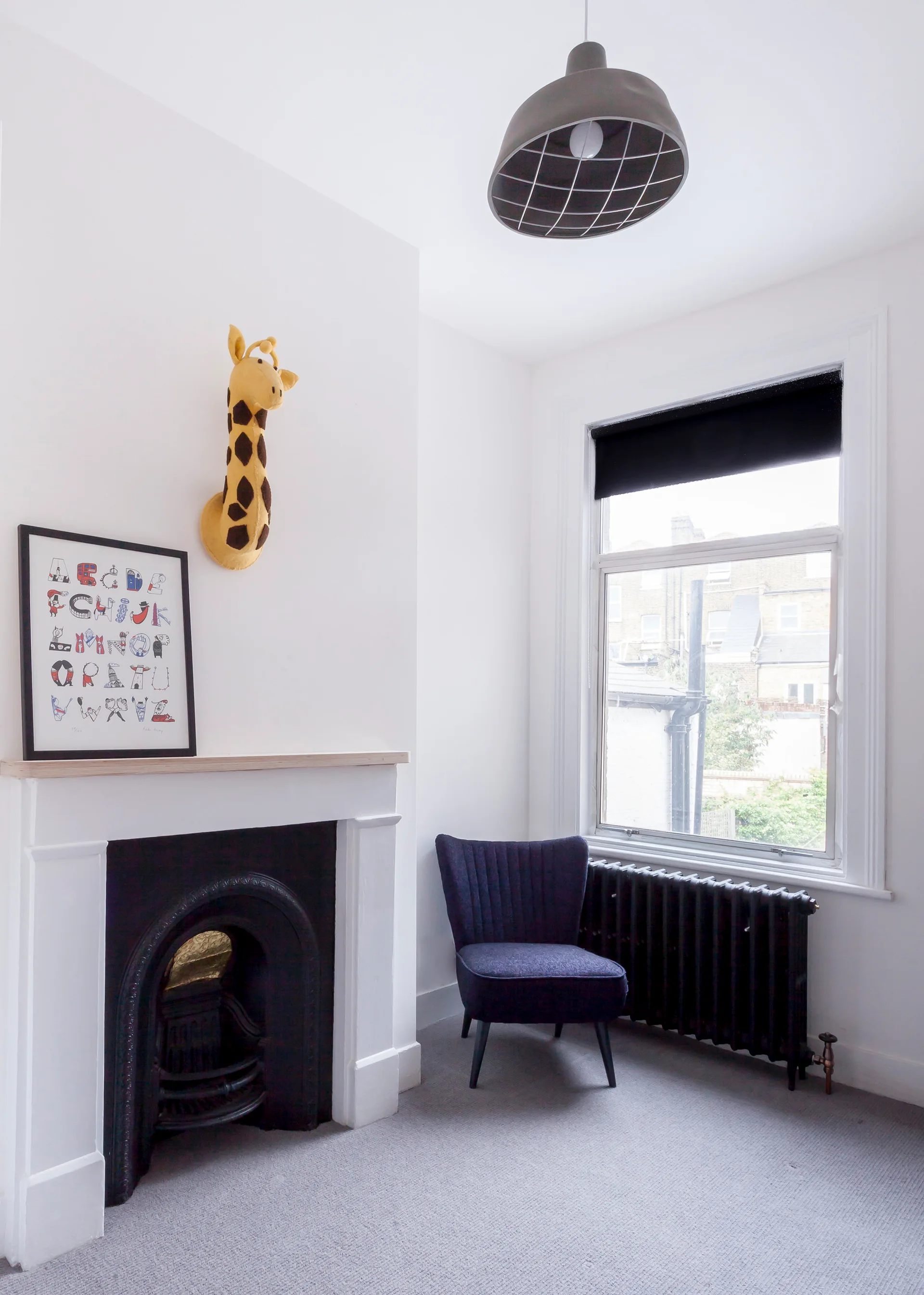 A white bedroom corner features a black cast iron fireplace, a blue velvet armchair, a black radiator, and a stuffed giraffe head wall mount.
