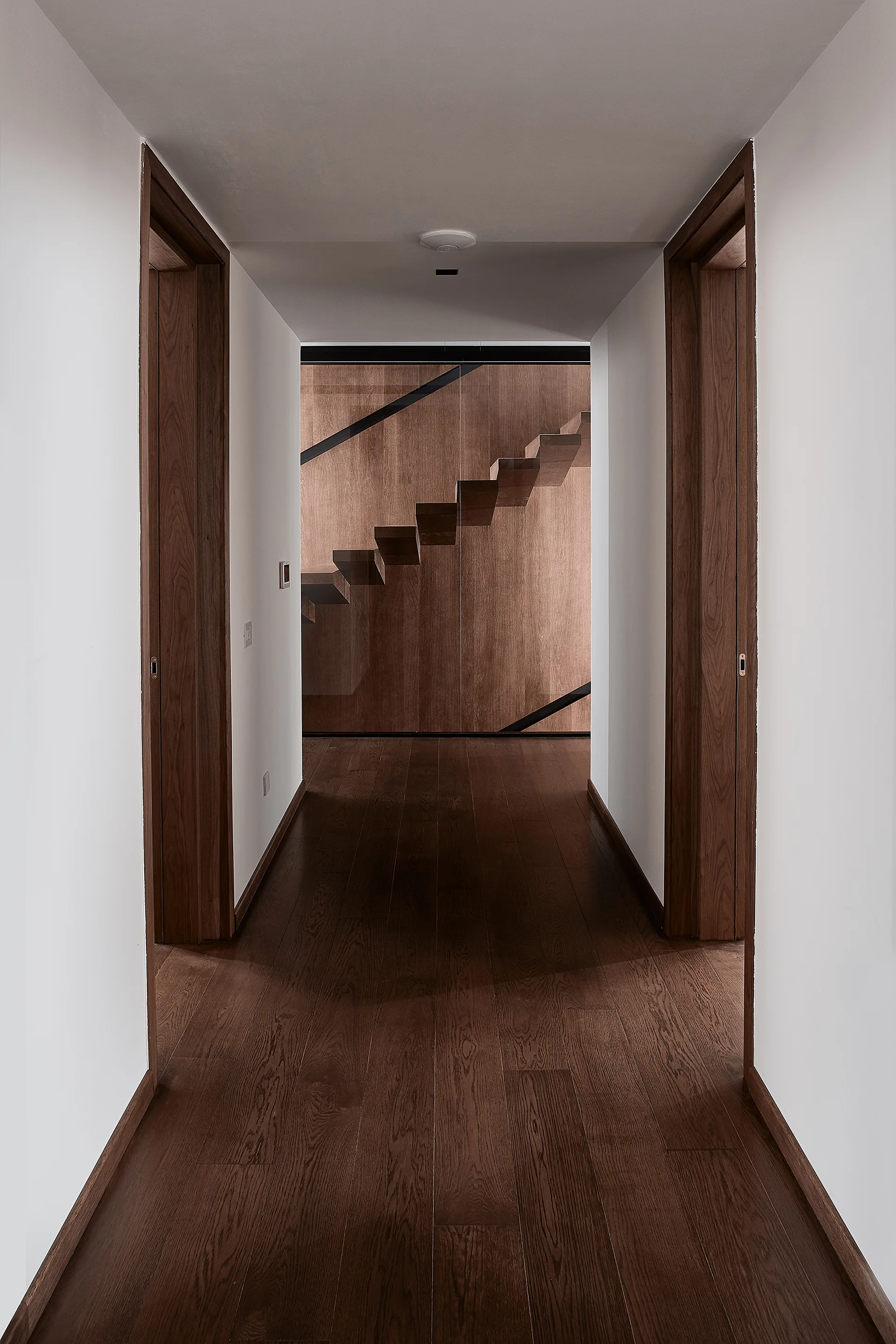 A perspective view down a minimalist white hallway with dark wood flooring and door frames, leading to a floating wooden staircase behind glass.
