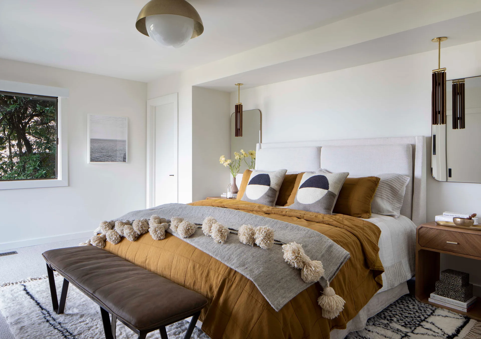 A modern bedroom features a bed dressed in gold-ochre bedding and a pom-pom throw blanket, flanked by wood nightstands and tall mirrors.