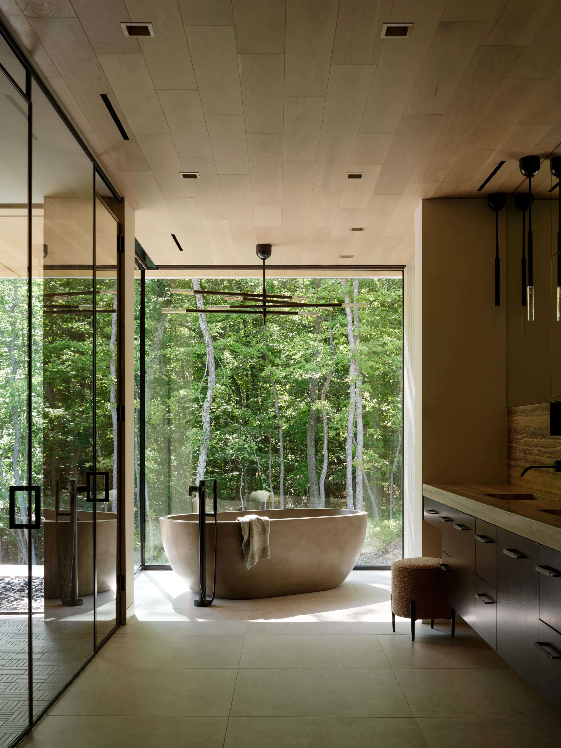 A modern bathroom features a freestanding stone bathtub positioned in front of a floor-to-ceiling window overlooking a green forest.