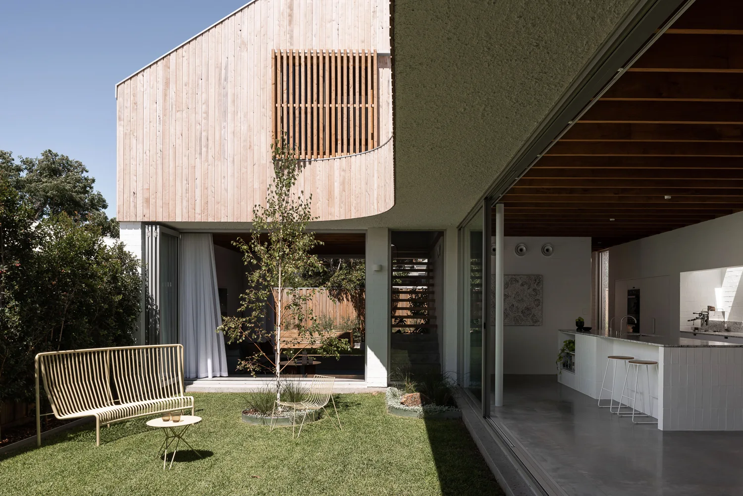 A modern wood-clad home features large sliding glass doors opening onto a grassy yard with yellow metal wire furniture and a view of the indoor kitchen.