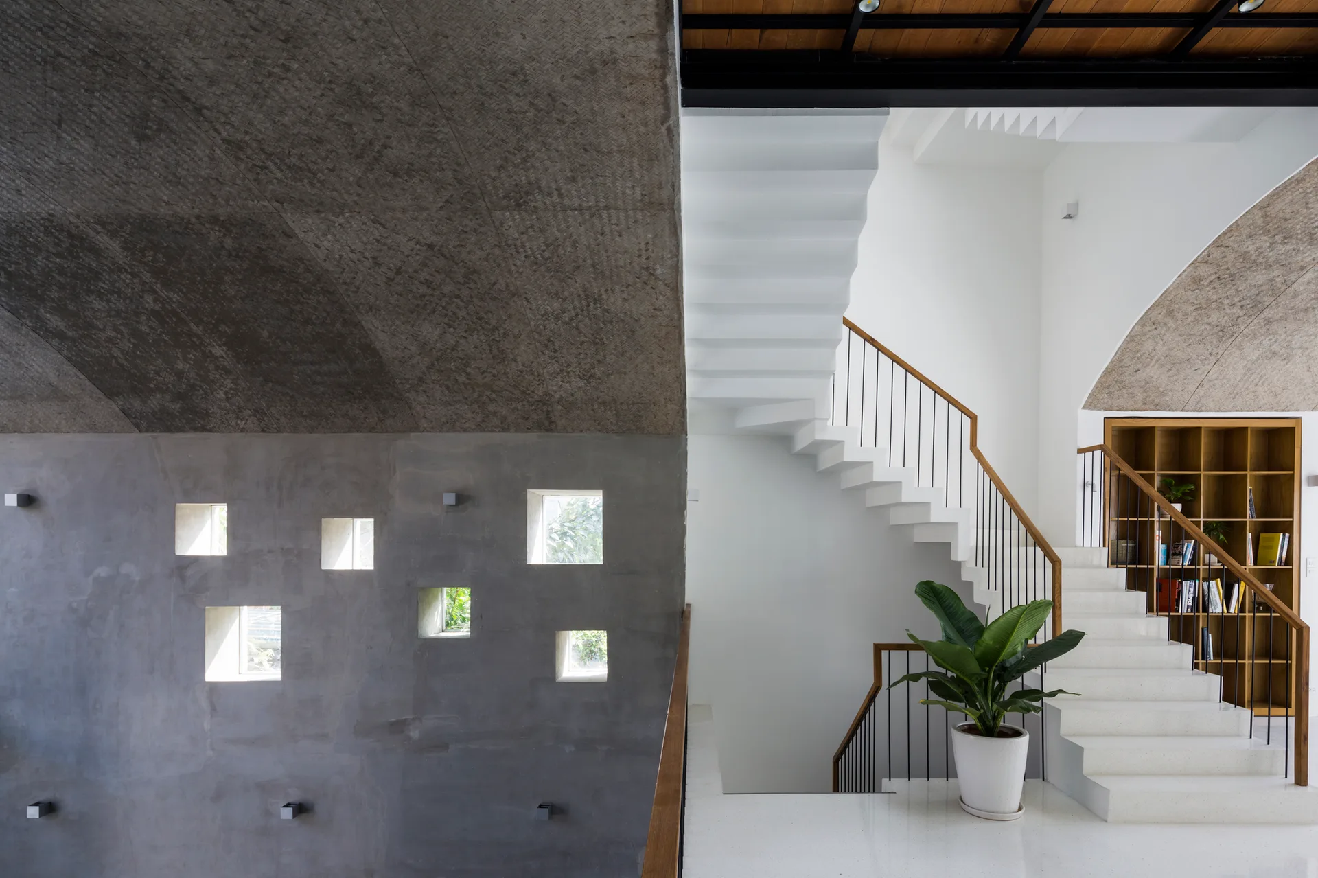 A white minimalist staircase with wooden handrails ascends next to a gray raw concrete wall featuring small square window cutouts.