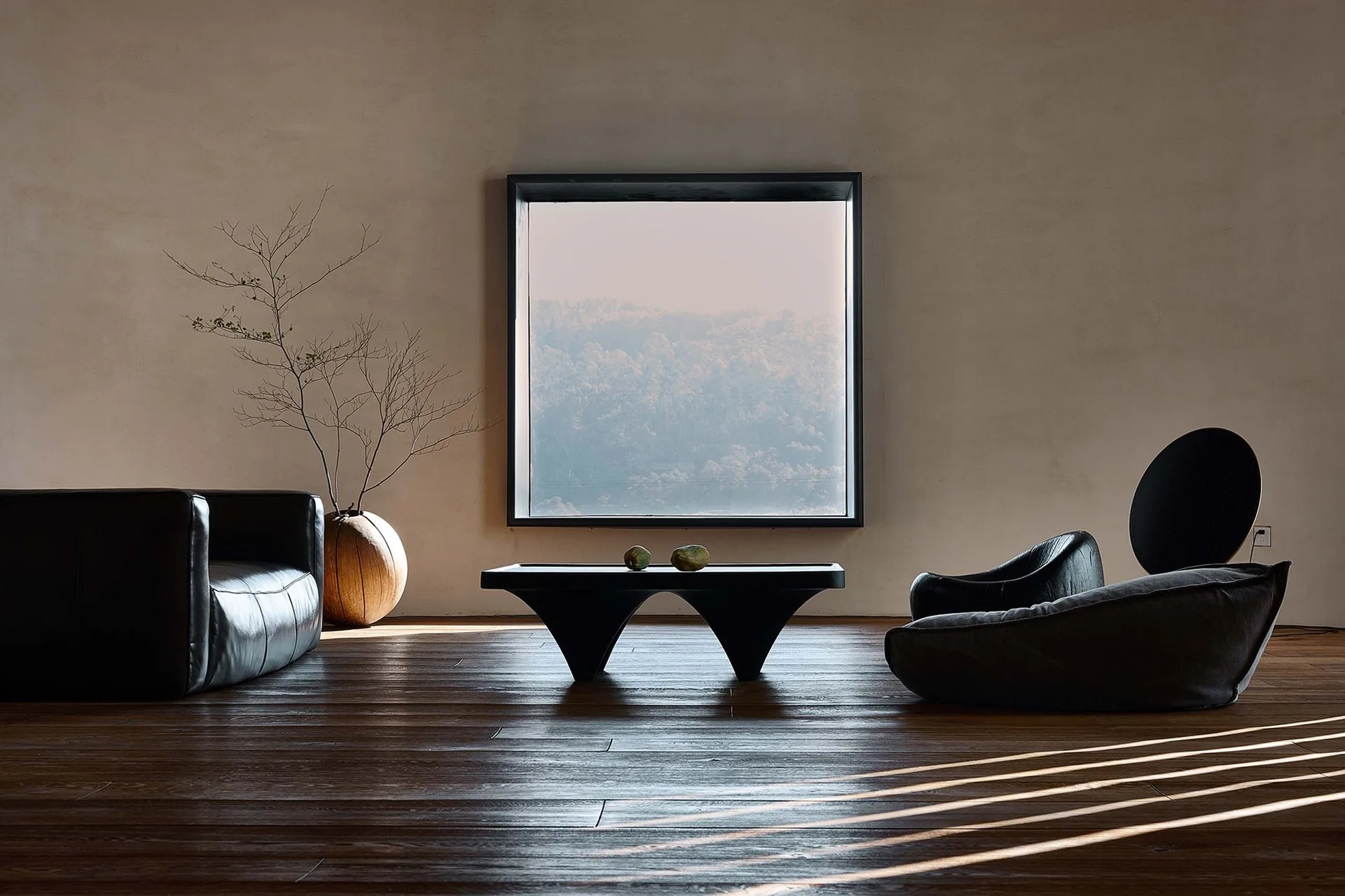 A minimalist living room with a large square window framing trees, low black leather and fabric seating, and a sculptural coffee table.