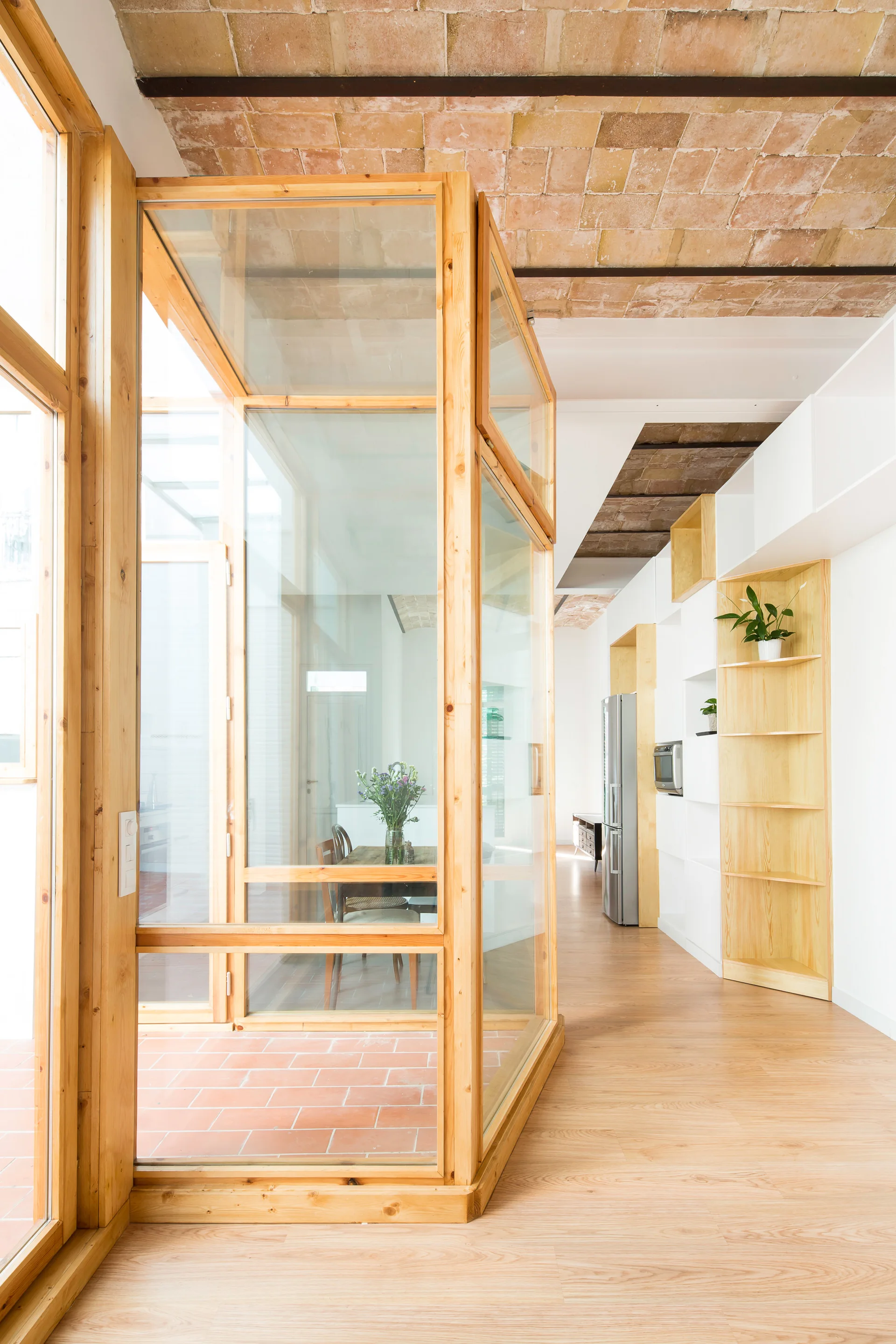 A light wood and glass partition stands on wood floors beneath a terracotta brick vaulted ceiling near a white kitchen storage unit.