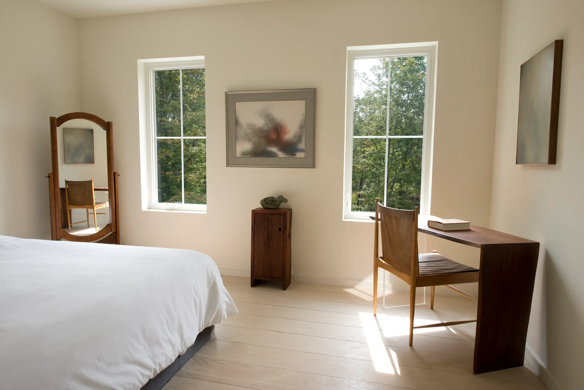 A minimalist bedroom features light wood floors, two windows overlooking trees, a wooden desk with a leather chair, and a freestanding mirror.