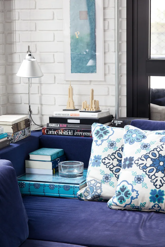 A close-up of a deep blue sofa styled with blue-and-white patterned pillows, stacks of books on the armrest, and a white brick wall backdrop.