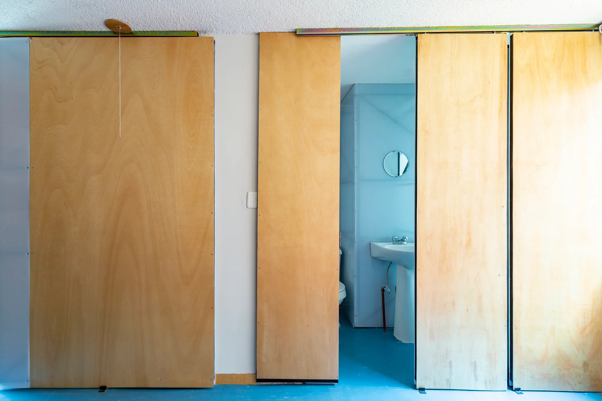 Large plywood sliding doors on a metal track open to reveal a bathroom with a white pedestal sink and a vibrant blue floor.