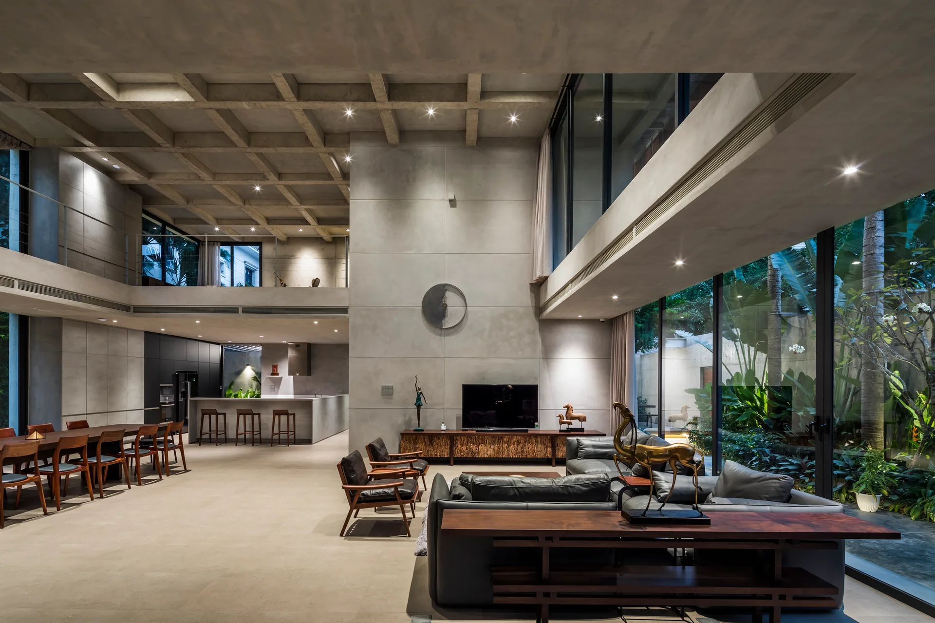 An open-concept living room with a concrete coffered ceiling, leather sofa, wood dining table, and massive glass walls showing outdoor greenery.